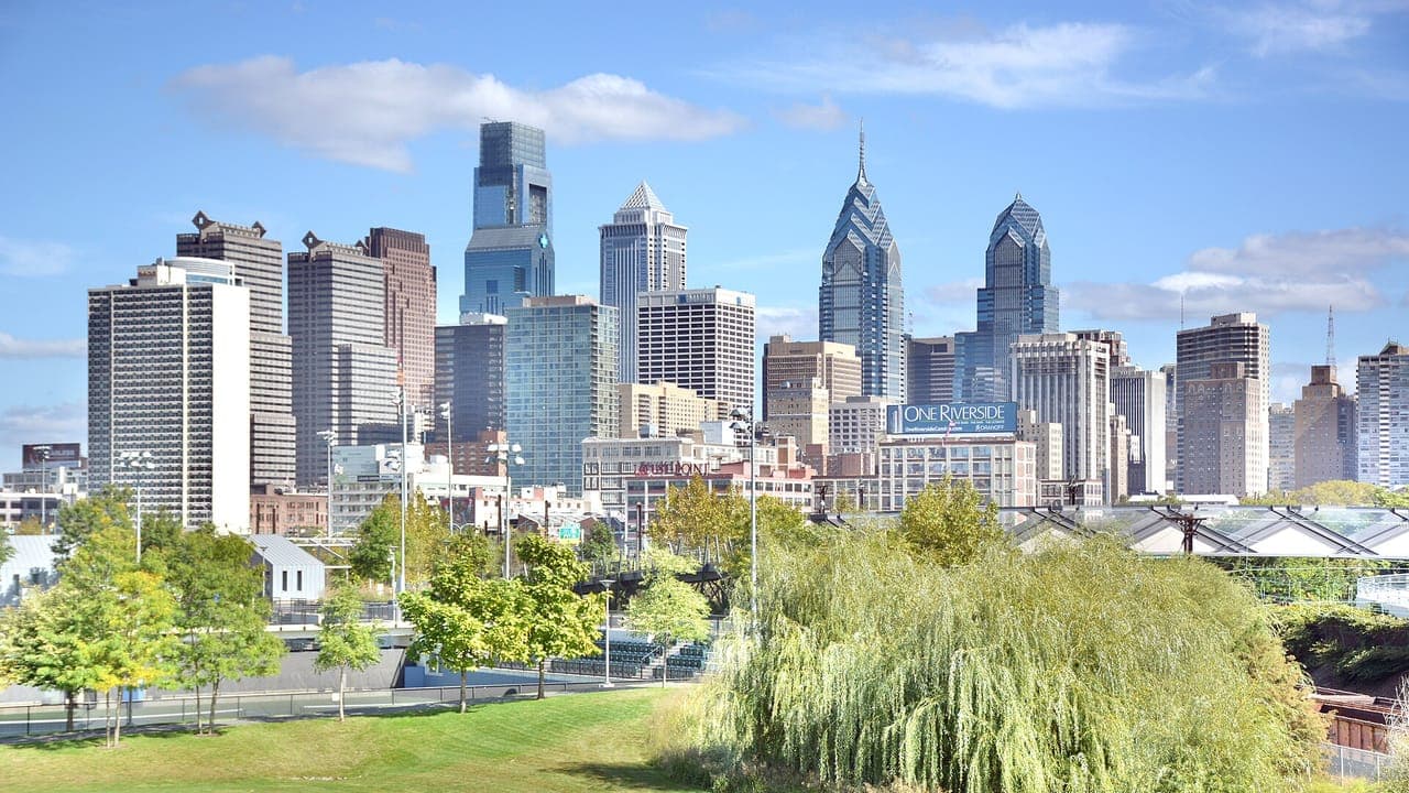Center City Philadelphia's glass and stone towers rise behind the lawn and trees of a riverside park on a sunny day