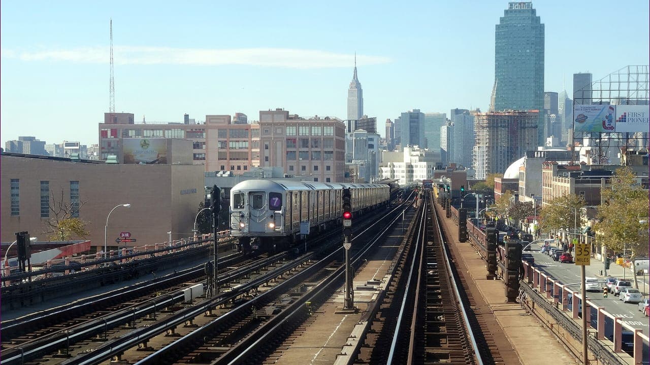 A silver 7 train runs on the elevated tracks above Queens Boulevard in Sunnyside, with Long Island City towers and the Empire State Building in the distance