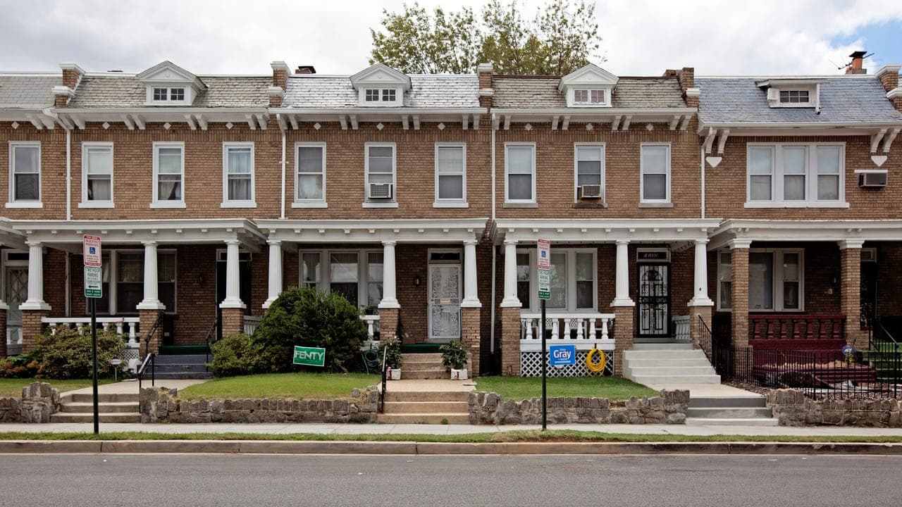 A row of two-story brick rowhouses with white-columned front porches and small front lawns on a street in Washington, DC