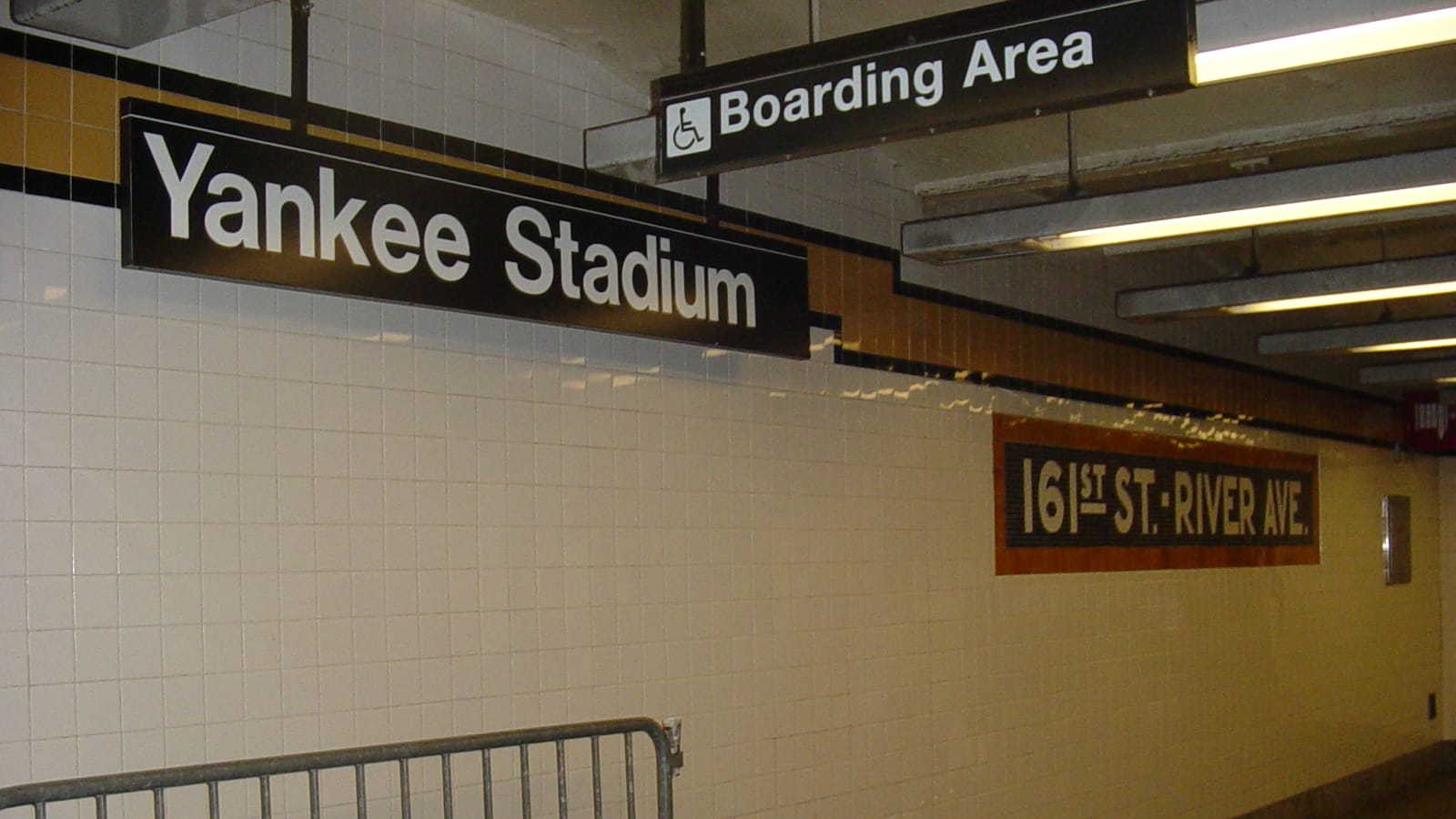 Photograph inside a subway station of a black sign reading Yankee Stadium, a Boarding Area sign with a wheelchair symbol, a white tiled wall with a mosaic reading 161st St.-River Ave., and a metal barrier in front