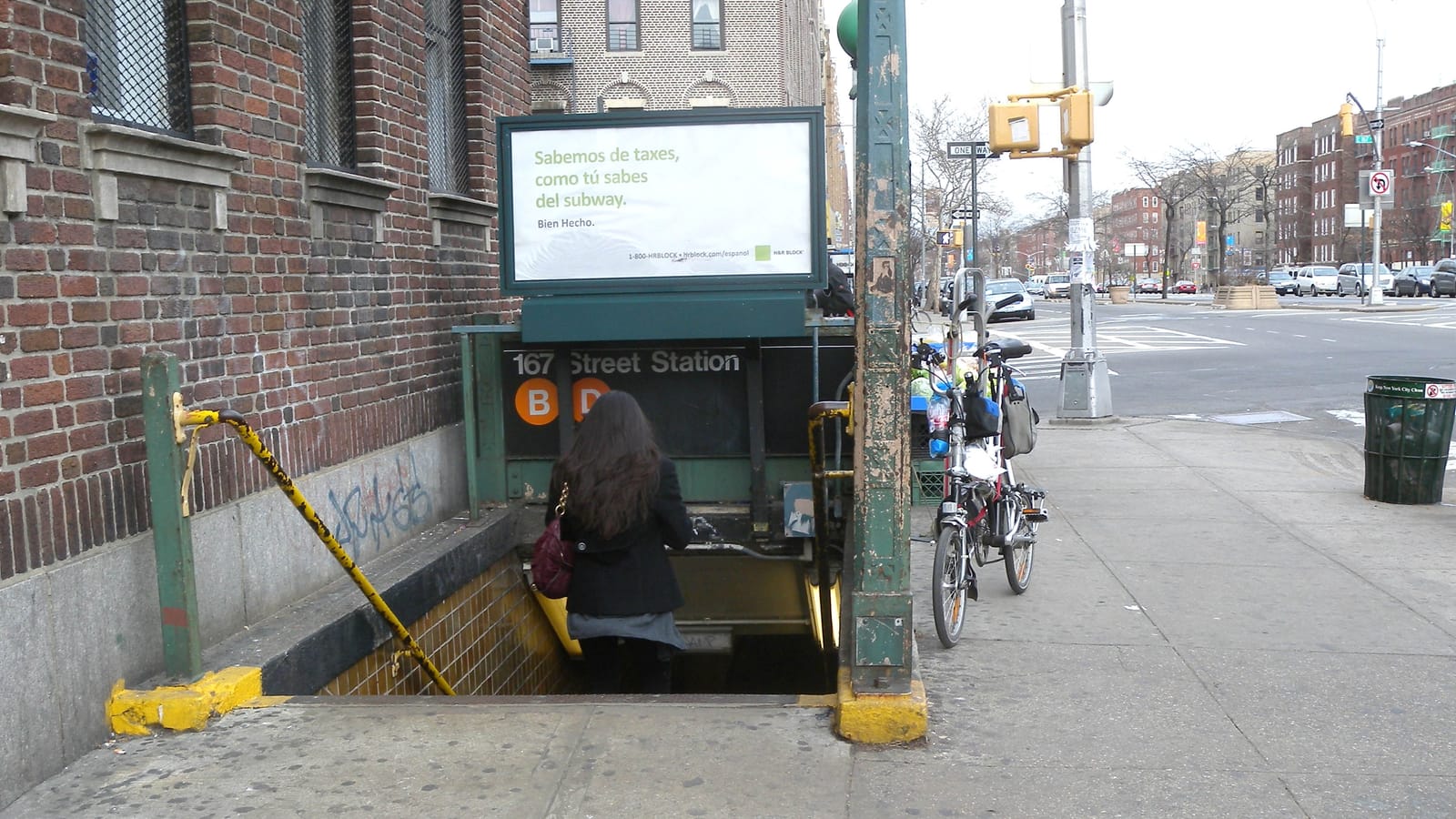 Photograph of a sidewalk subway stair entrance with a green railing and a sign reading 167 Street Station with orange B and D circles, a woman seen from behind walking down, a parked bicycle, a Spanish-language billboard above and apartment buildings along a wide avenue