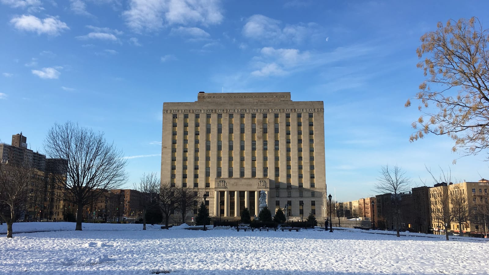 Photograph of a snow-covered park lawn with bare trees in front of a tall pale stone courthouse building with columns at its base, under a blue sky with clouds, and apartment buildings at both sides