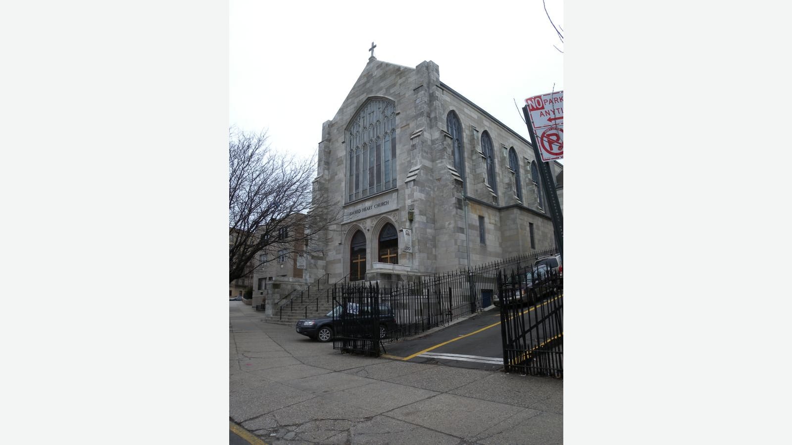Photograph of a gray stone Gothic-style church with a tall pointed window, a cross on the roof and the words Sacred Heart Church over two arched doors, behind a black iron fence, with a parked car, a sloping driveway and a no-parking sign, shown on a plain light background