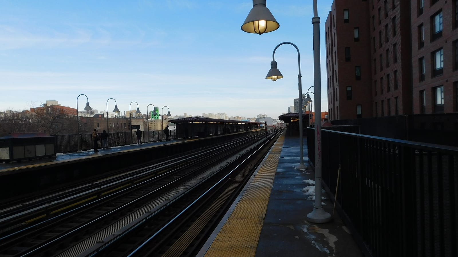 Photograph of an elevated subway platform on a clear winter day, with curved gray lamp posts, a yellow tactile strip along the platform edge, tracks running to a far canopy, a few people waiting on the opposite platform and tall brick apartment buildings at right