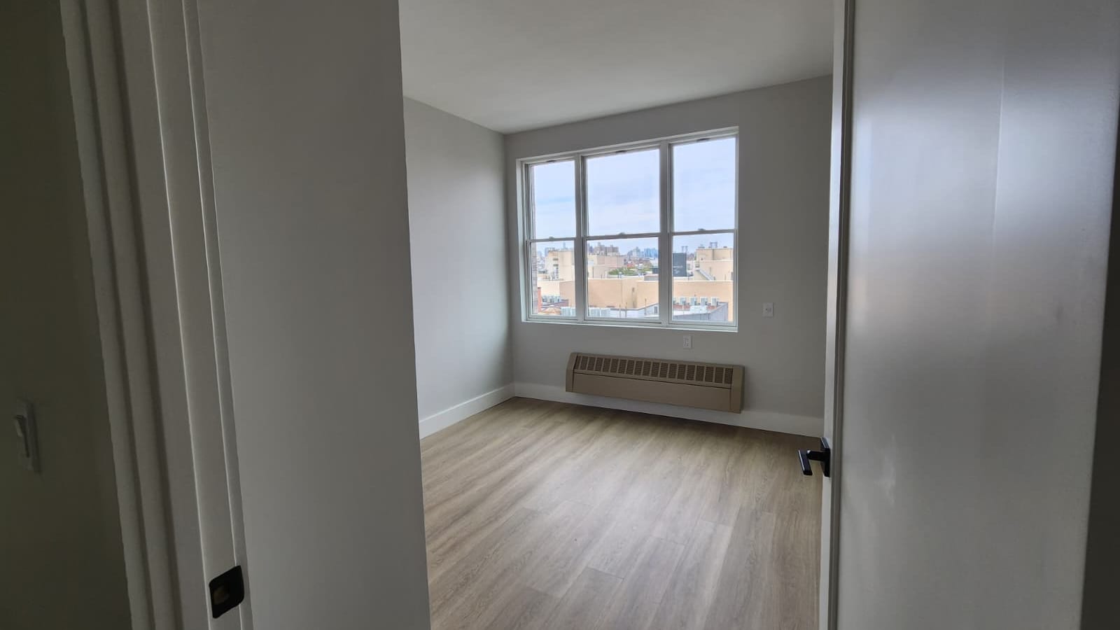 Photograph through a doorway of an empty room with pale grey walls, wood-look plank flooring, a three-part window looking out over rooftops and a covered unit mounted on the wall below the window
