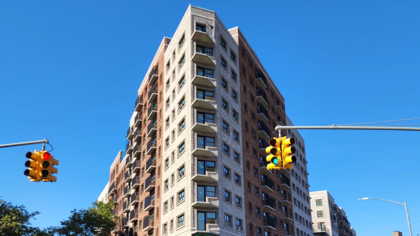 Photograph looking up at the pointed corner of a tall apartment building in pale grey and red brick with balconies at each floor, against a clear blue sky, with two yellow traffic signals in the foreground and treetops at the lower left