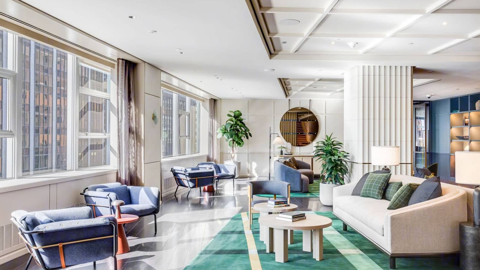 Photograph of a long sunlit lounge with blue armchairs along tall windows, a cream curved sofa with green plaid cushions, round wooden coffee tables on a green striped rug, potted plants, a fluted white column and a coffered ceiling