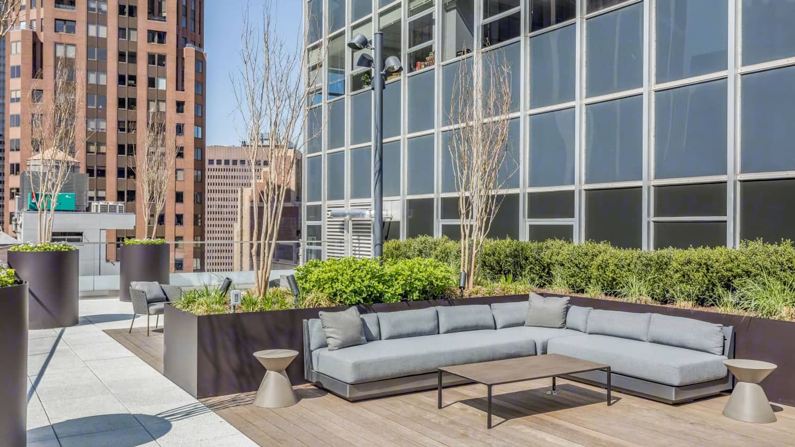 Photograph of a roof terrace with a grey L-shaped outdoor sofa, a low table and two small side tables on a wood deck, planters with shrubs and bare young trees, a glass and metal wall of the tower rising behind, and a brick high-rise across the street
