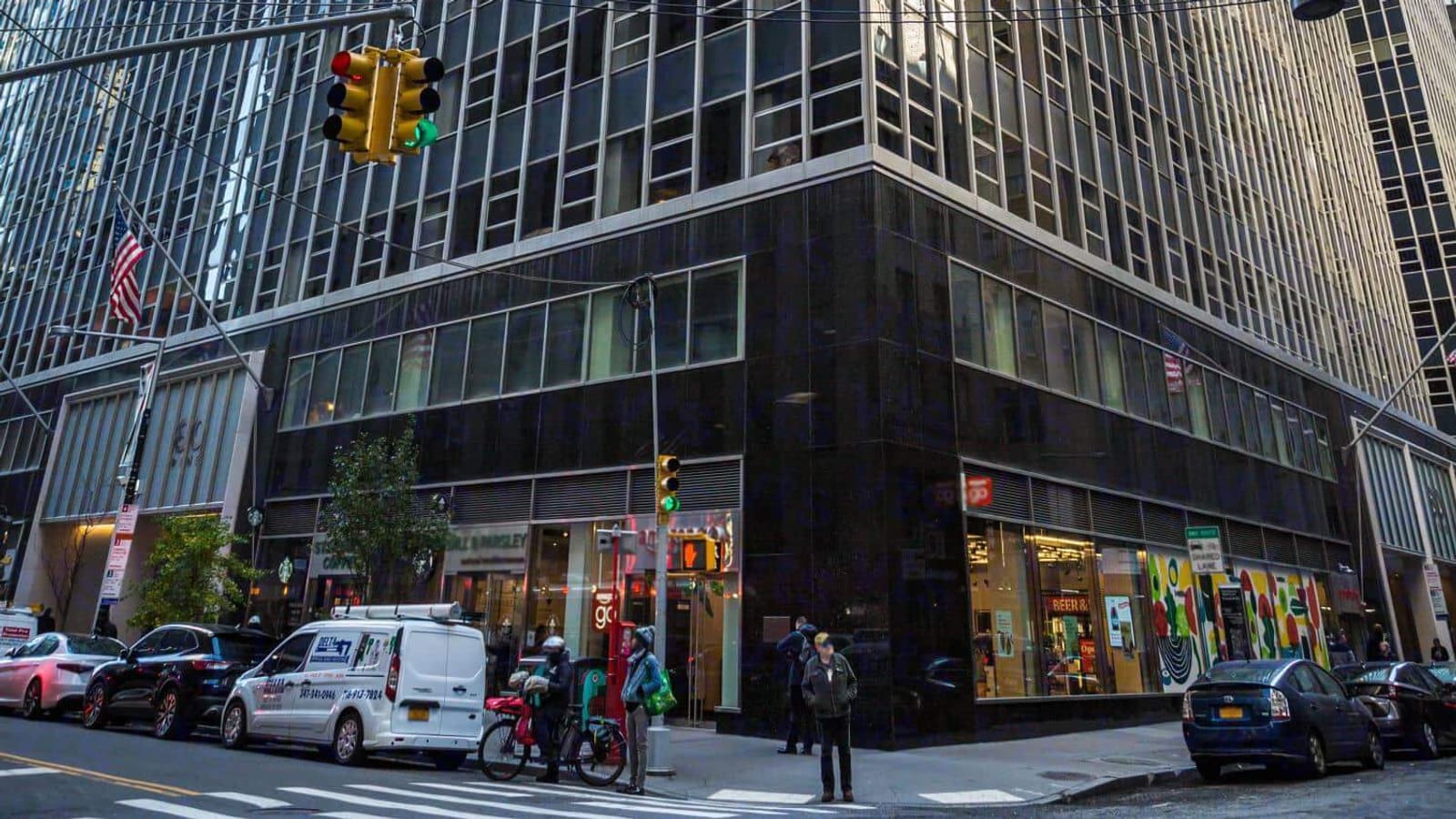 Photograph of the lower floors of a glass and metal office tower on a street corner, with a polished black stone base, ground-floor shop windows, a traffic light and an American flag on the facade, parked cars and a white van at the curb, and a few people waiting at the crosswalk