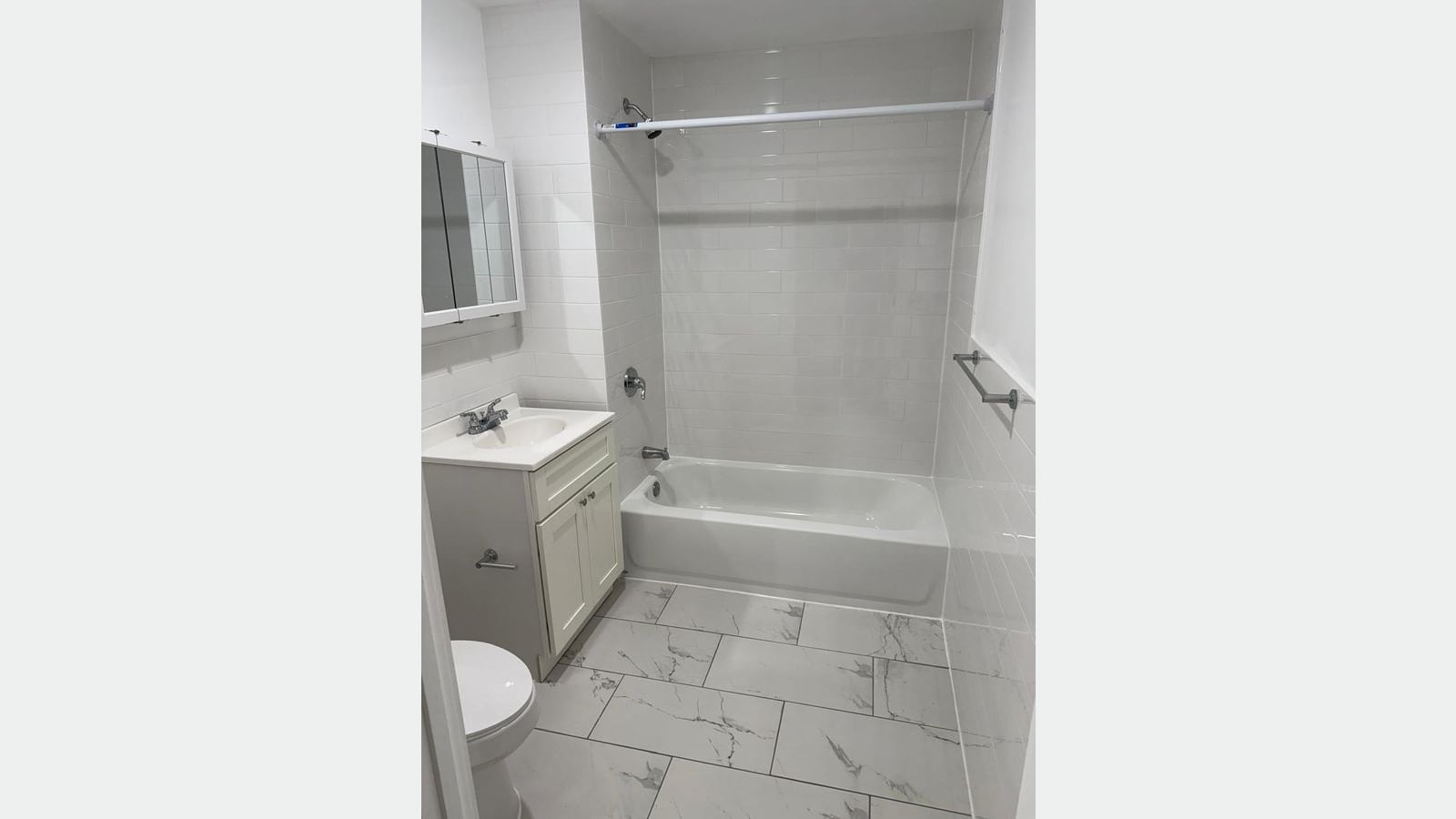 Photograph of a bathroom with white subway tile walls, a white tub with a shower rod, a white vanity with a mirrored cabinet above it, a toilet and large gray-veined white floor tiles
