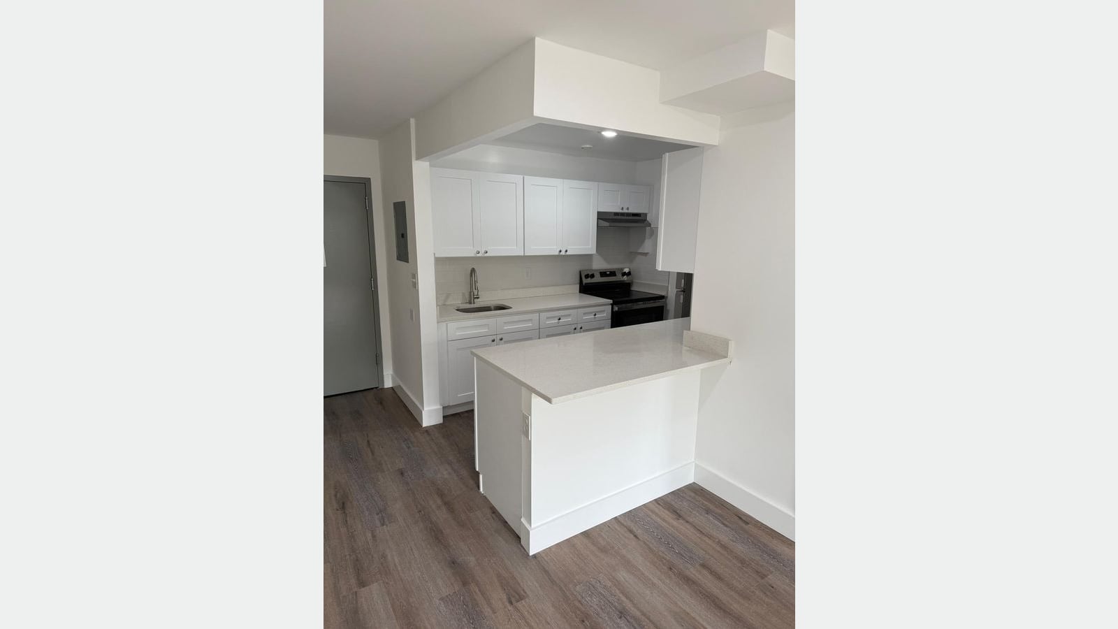 Photograph of a small open kitchen with white shaker cabinets, a white stone counter and peninsula, a stainless steel sink, a stainless steel electric range with a range hood, gray wood-look flooring and a gray apartment door at left