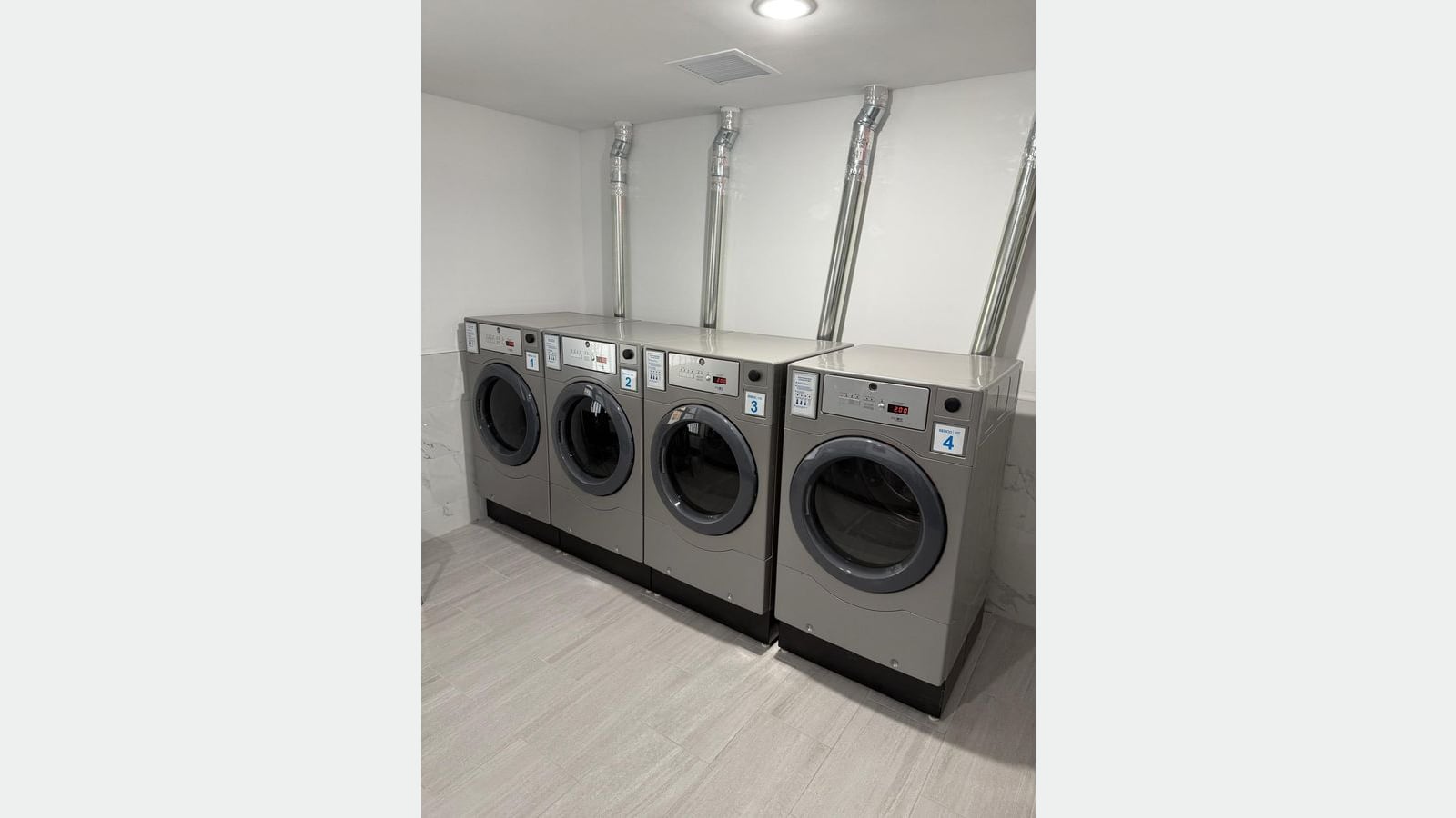 Photograph of a laundry room with four gray front-loading machines numbered 1 to 4 against a white wall, metal vent pipes above them and light gray floor tiles
