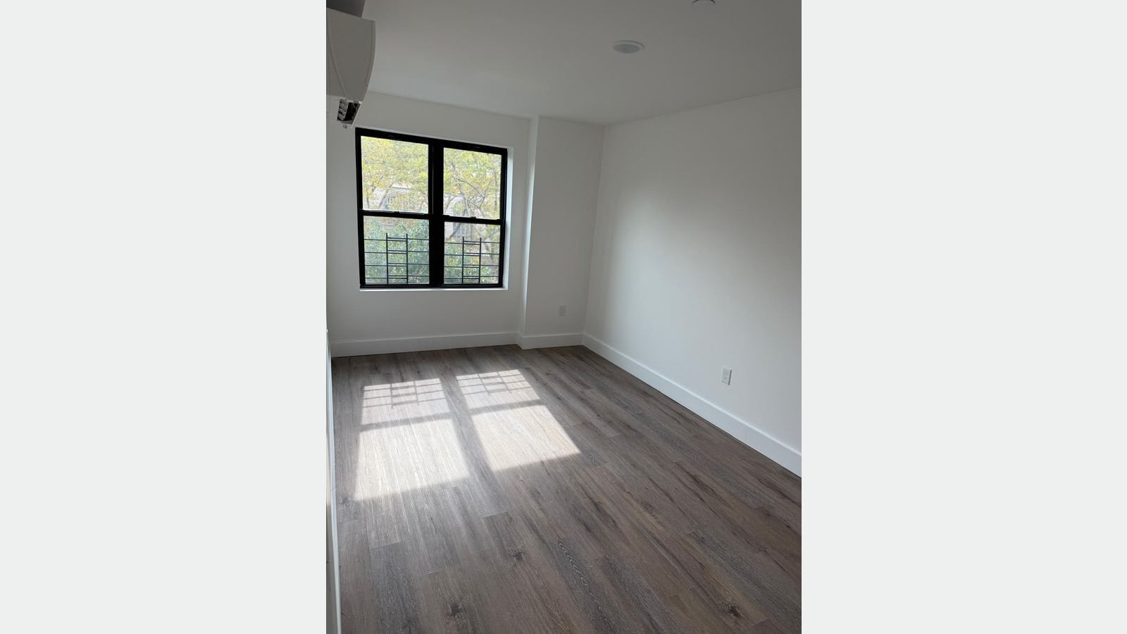 Photograph of an empty room with white walls, gray wood-look plank flooring, sunlight on the floor, a black-framed double window with trees outside and a wall-mounted heating and cooling unit near the ceiling