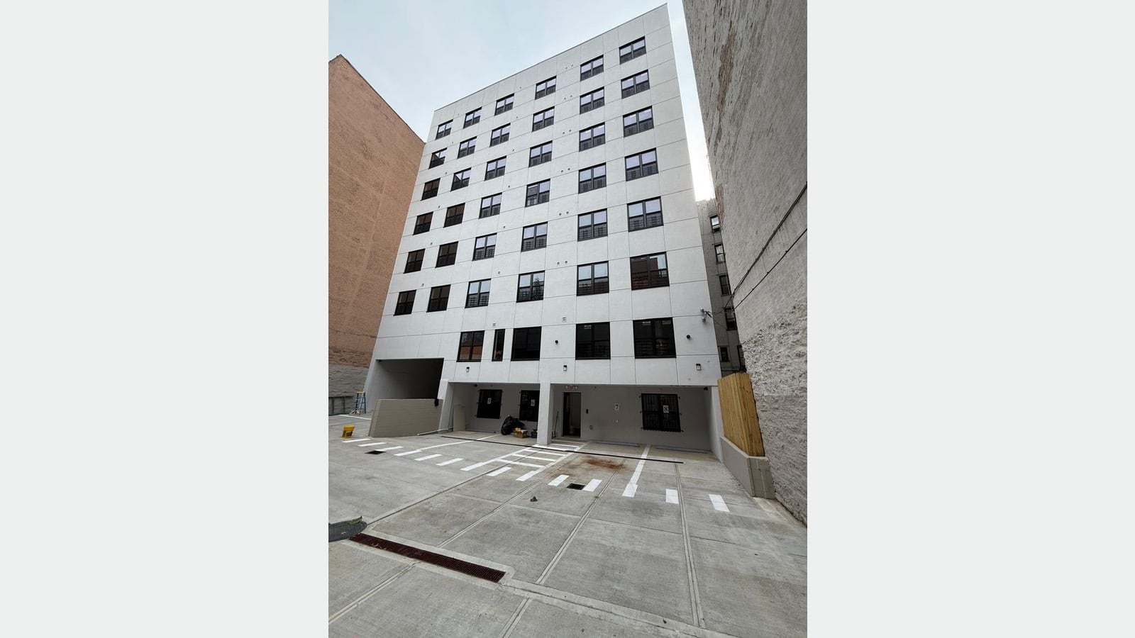 Photograph of the rear of a white seven-story apartment building with rows of black-framed windows, a ground level set back over a paved lot with painted parking lines, and brick walls of older buildings on both sides
