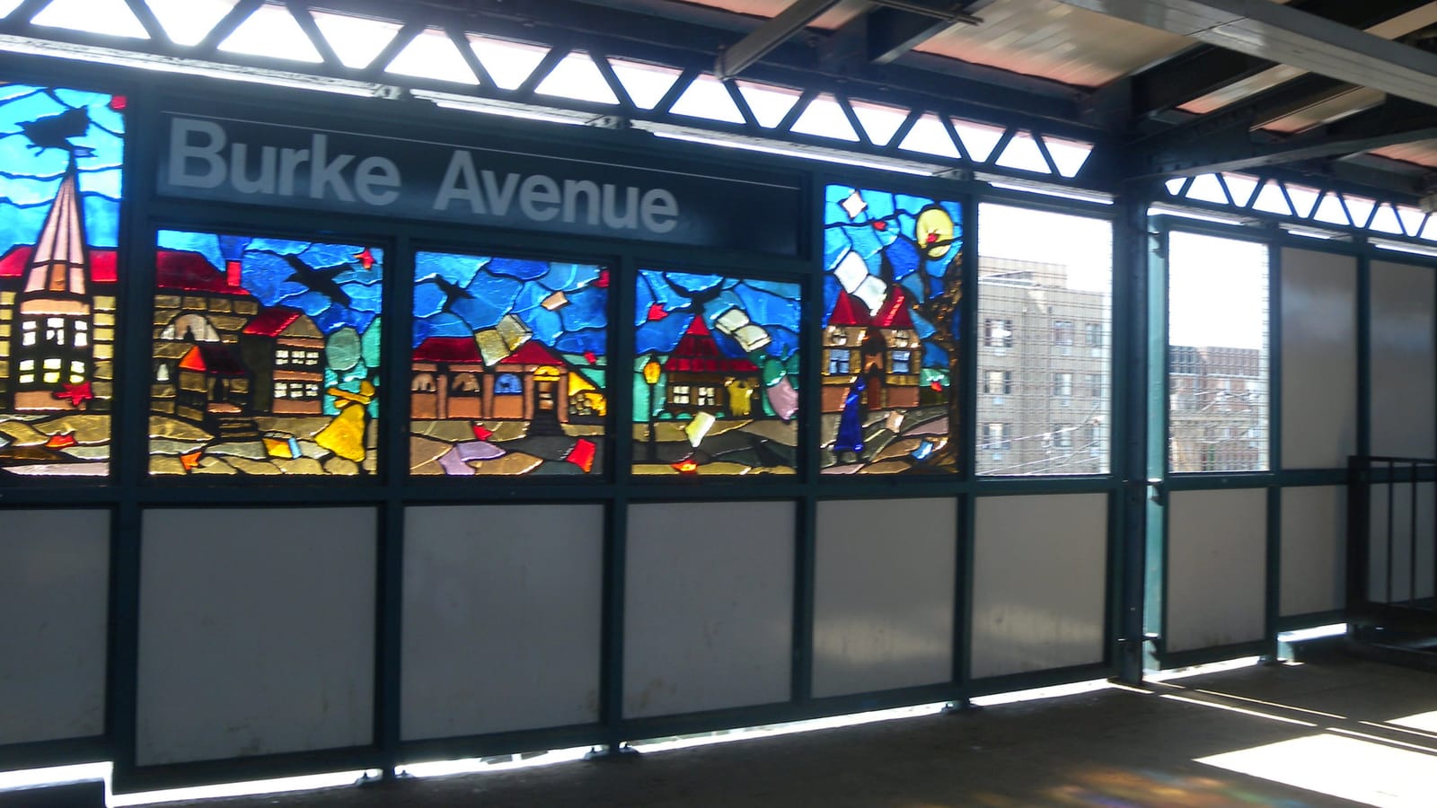 Photograph of an elevated subway platform wall with a sign reading Burke Avenue above colorful stained-glass panels showing houses, a church steeple, birds and flying pages against a blue sky, with sunlight casting colored light on the platform floor and apartment buildings visible through the windows at right