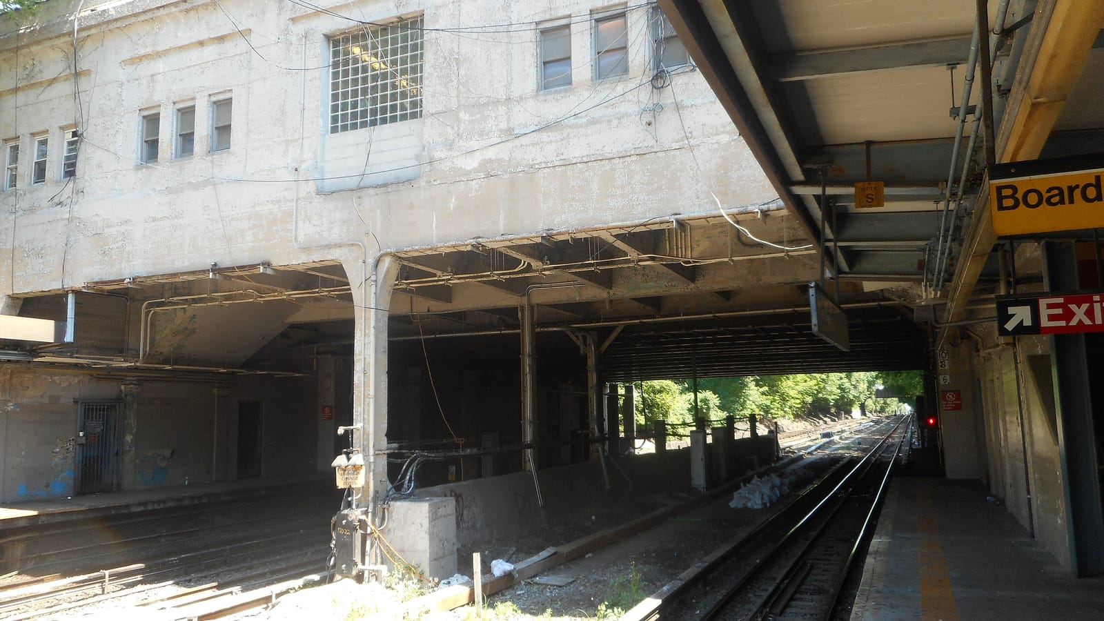 Photograph from a train platform of a weathered concrete station house built over the tracks on thick columns, with small windows and a glass-block window, two tracks running under it toward trees in sunlight, and a yellow sign reading Board and a red Exit sign on the platform at right