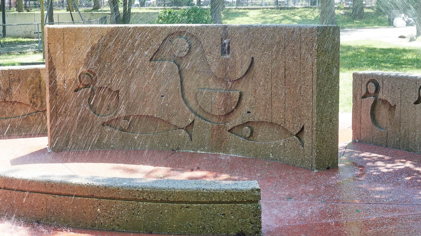 Photograph of curved concrete walls in a park spray area, carved with simple shapes of a large bird, ducks and fish, with water spraying across the red ground, and grass, trees and a fence behind
