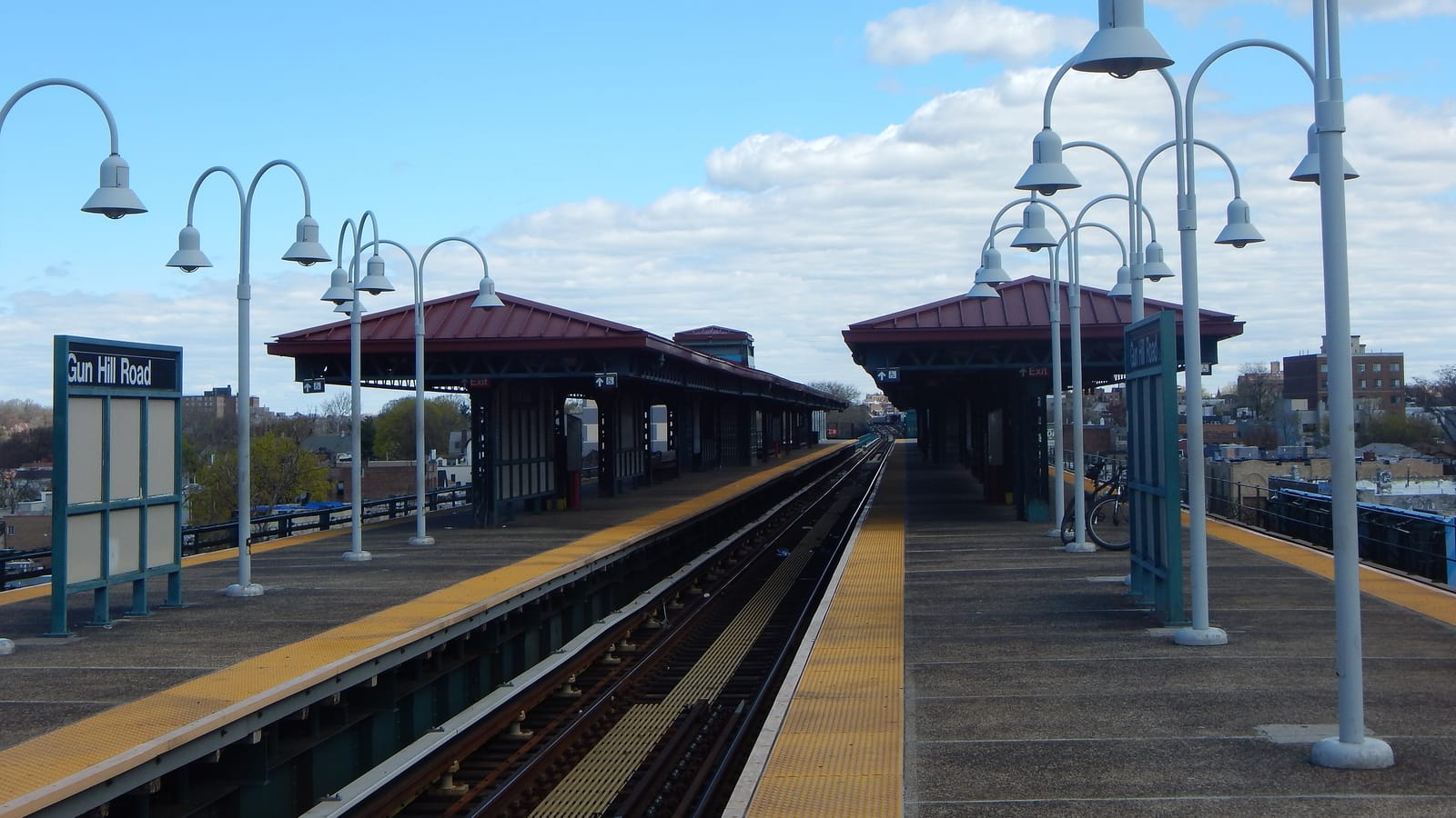Photograph of an elevated subway station with two empty platforms on either side of the tracks, red-roofed shelters, rows of curved white lamp posts, a green sign reading Gun Hill Road, and low buildings and trees below under a partly cloudy sky