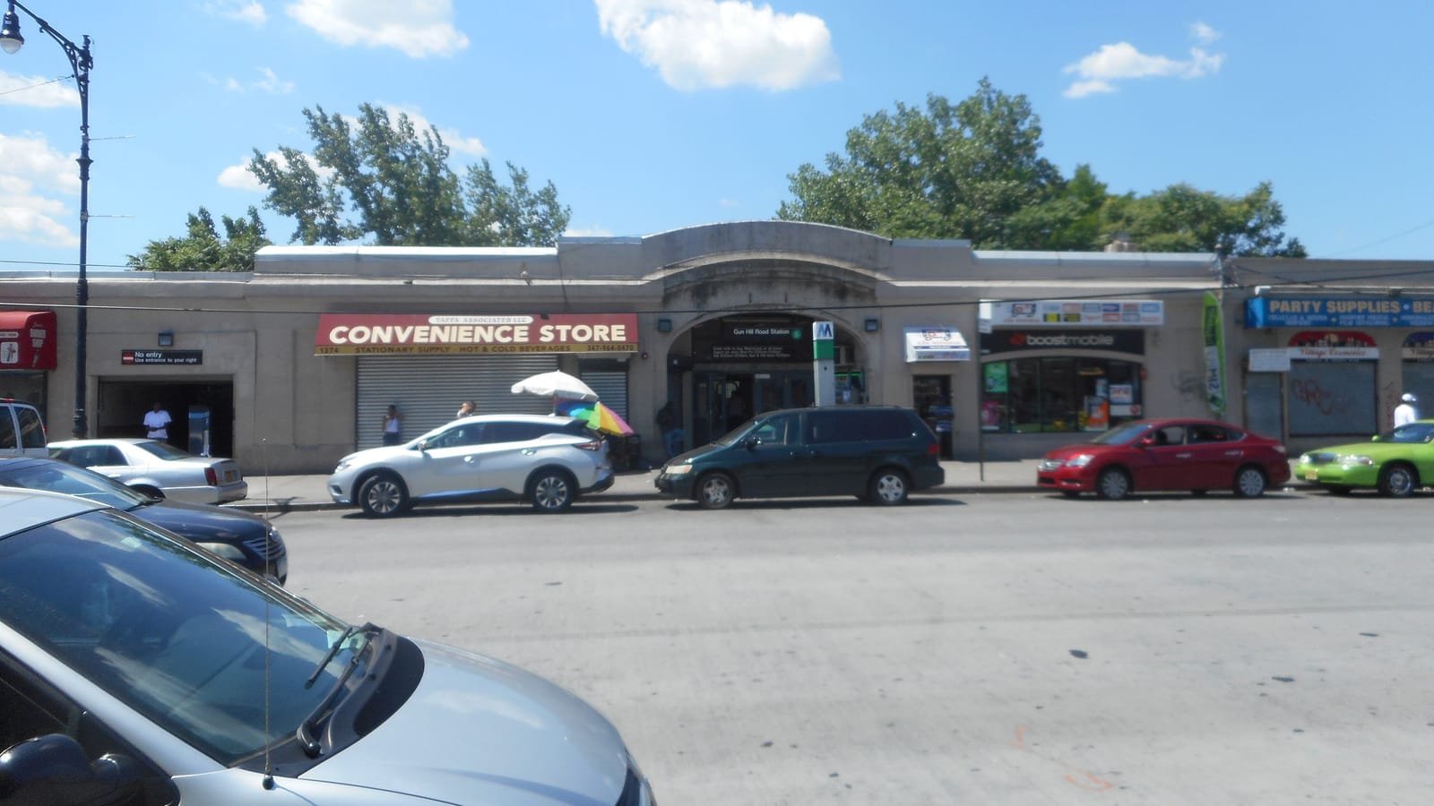Photograph of a long, low, pale stone station house on a wide street under a blue sky, with an arched entrance in the middle under a sign for Gun Hill Road Station and the 5 train, a green M sign post, shops on either side including a convenience store with a red awning and a phone store, parked cars along the curb and the hood of a silver car in the foreground