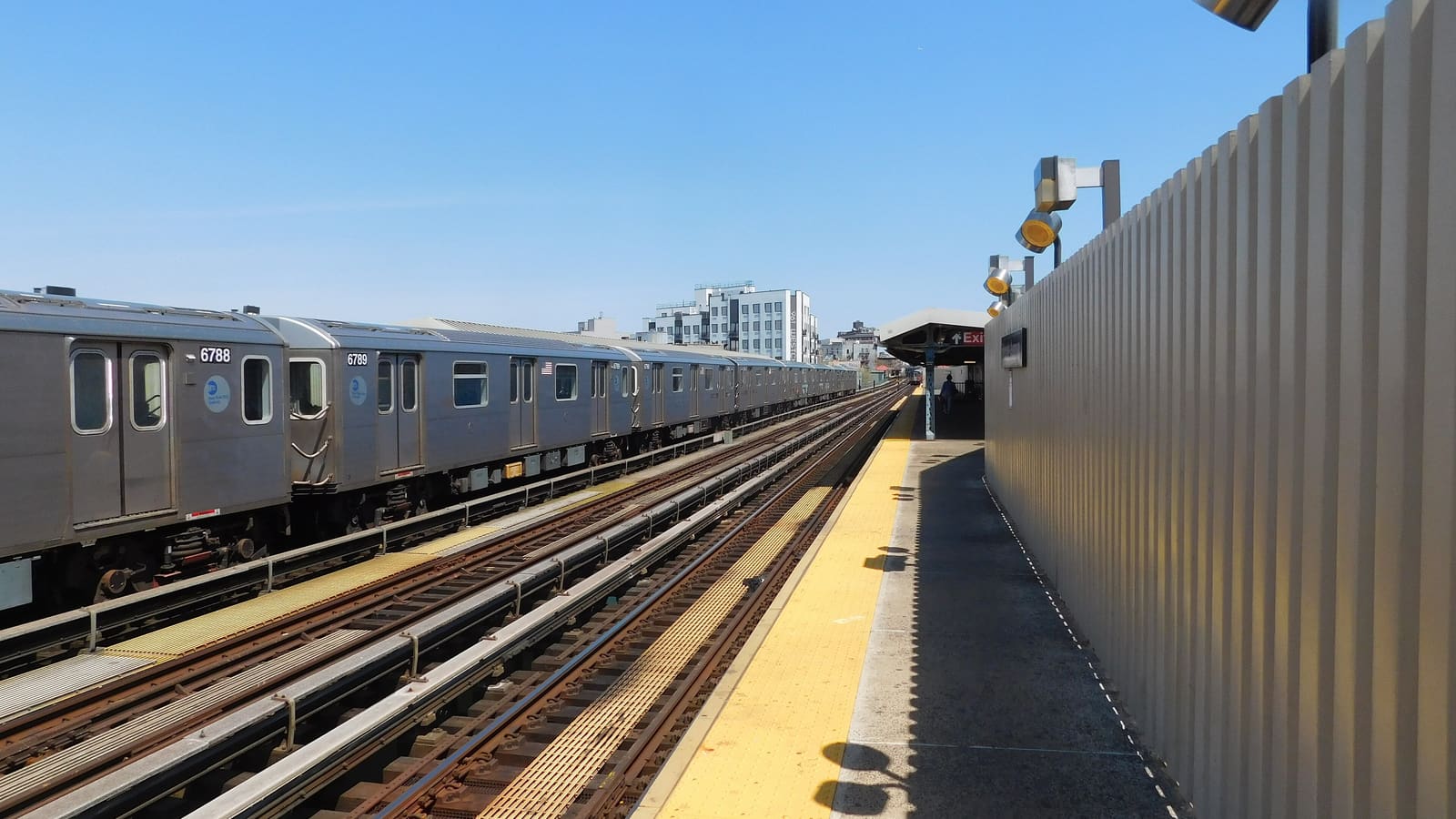 An open-air subway platform with a yellow tactile edge strip, a silver train on the far track, a ribbed grey windbreak wall at right and a red Exit sign under a canopy at the far end, under a clear blue sky