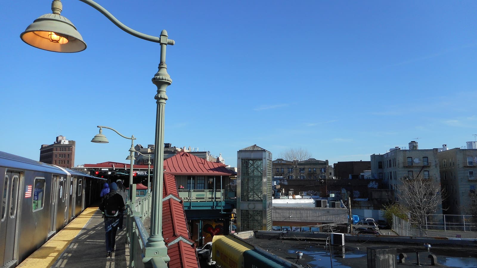 A subway platform with green lamp posts and a silver train at left, a red-roofed station house below, and a glass elevator tower rising beside apartment buildings under a blue sky
