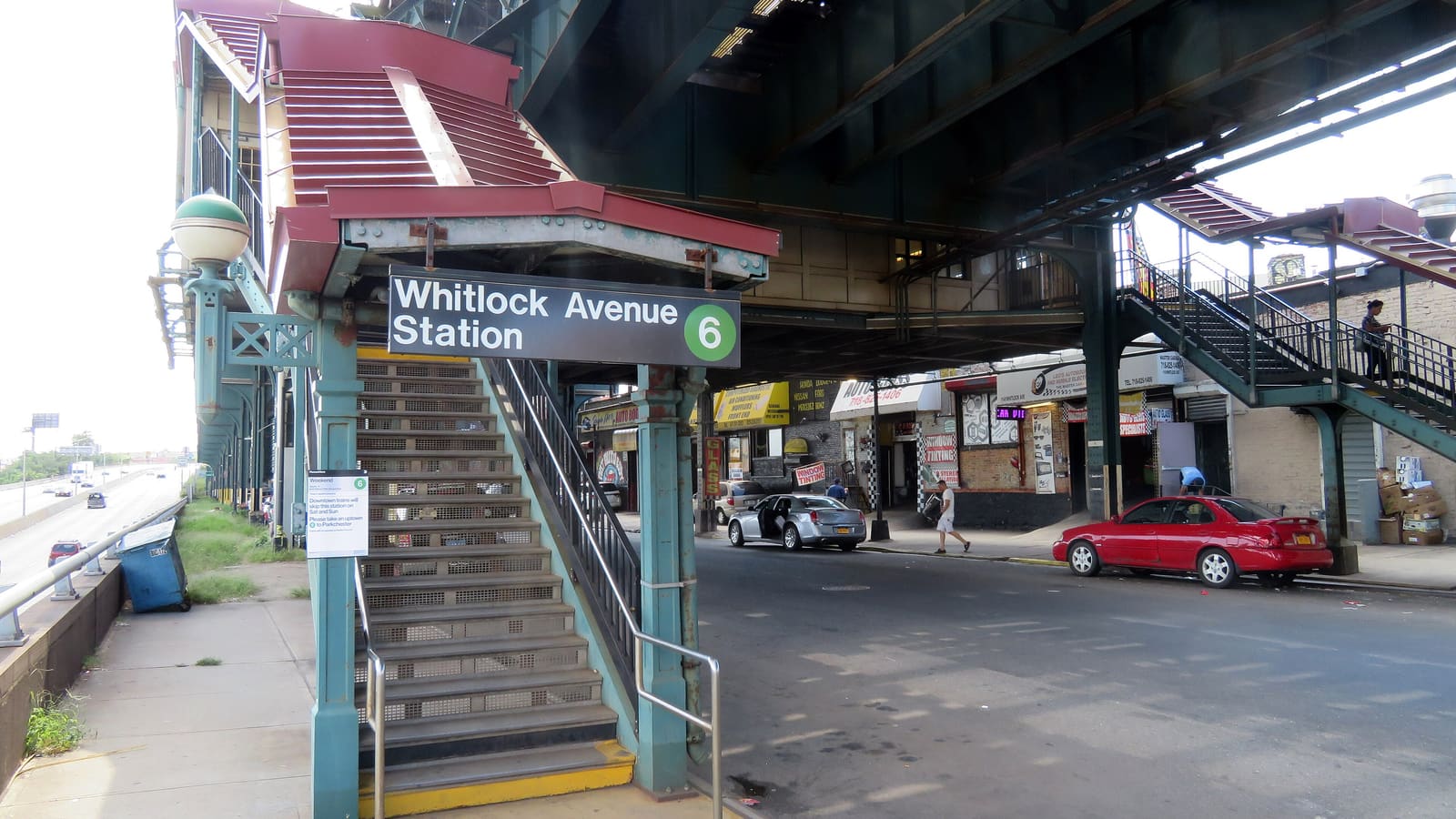 Green steel street stairs with a red canopy and a sign reading Whitlock Avenue Station 6, under a steel subway structure over a street lined with auto shops, with a red car and a silver car parked at the curb