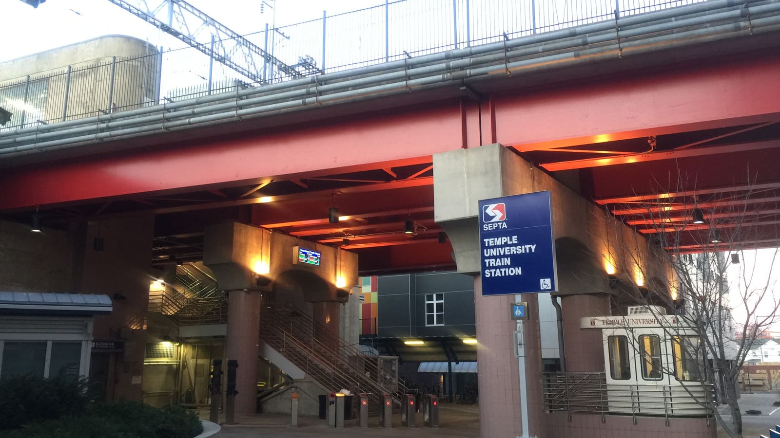 Street-level entrance beneath a red steel railroad viaduct with a blue SEPTA sign reading Temple University Train Station and stairs leading up to the platforms