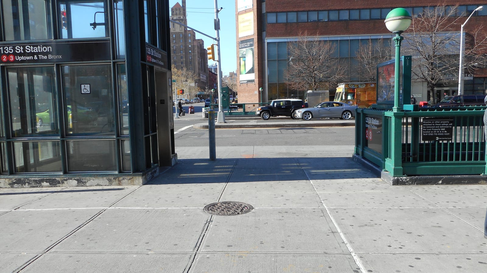 Photograph of a glass subway elevator booth with a wheelchair symbol and a sign reading 135 St Station, 2 3, Uptown and The Bronx, beside a green railed stair entrance, a wide sidewalk and a street with cars and a brick building behind