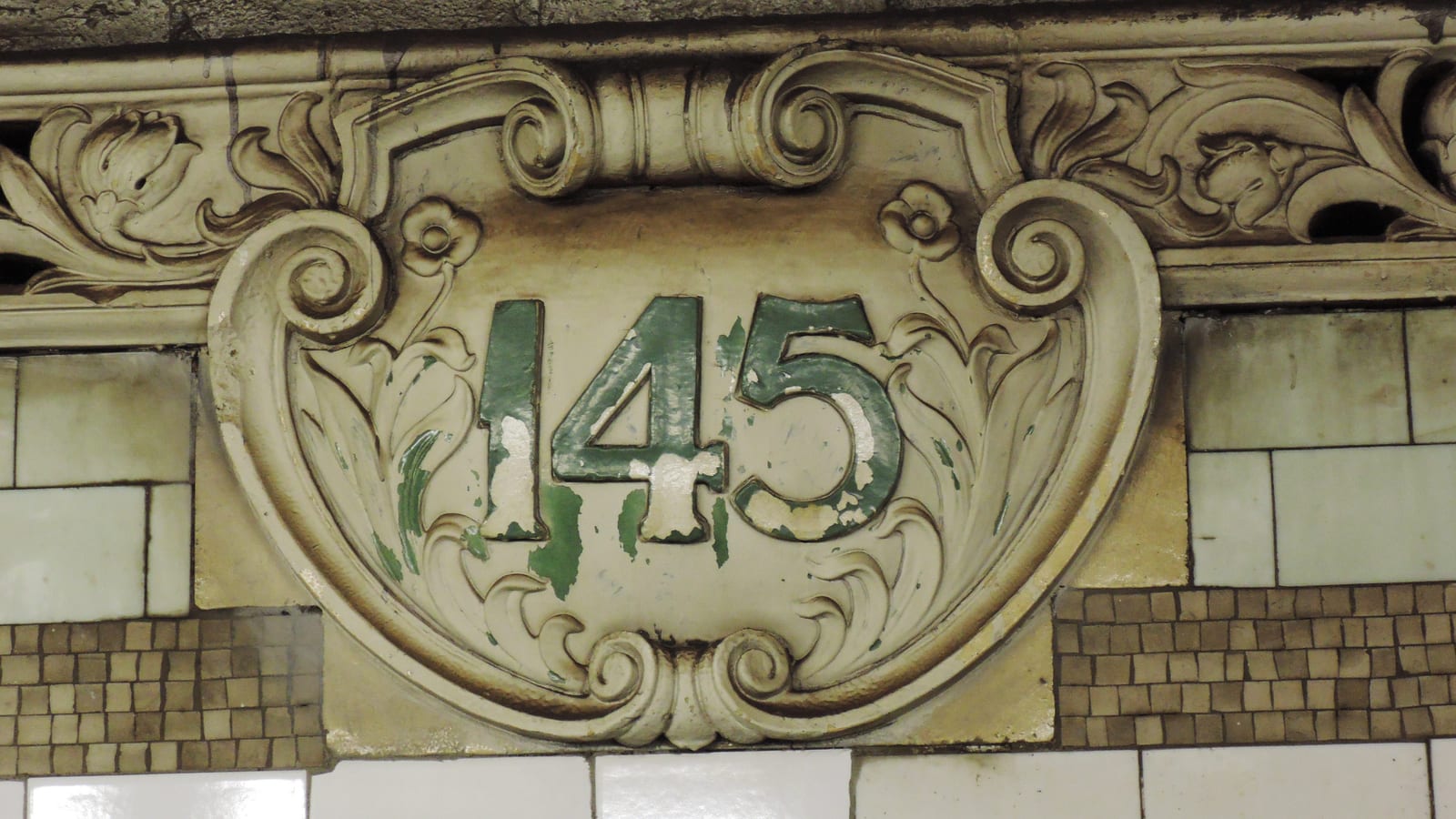 Photograph of a cream-colored ornamental plaque on a subway wall with the number 145 in chipped green paint, framed by carved scrolls and flowers, above white and brown tiles