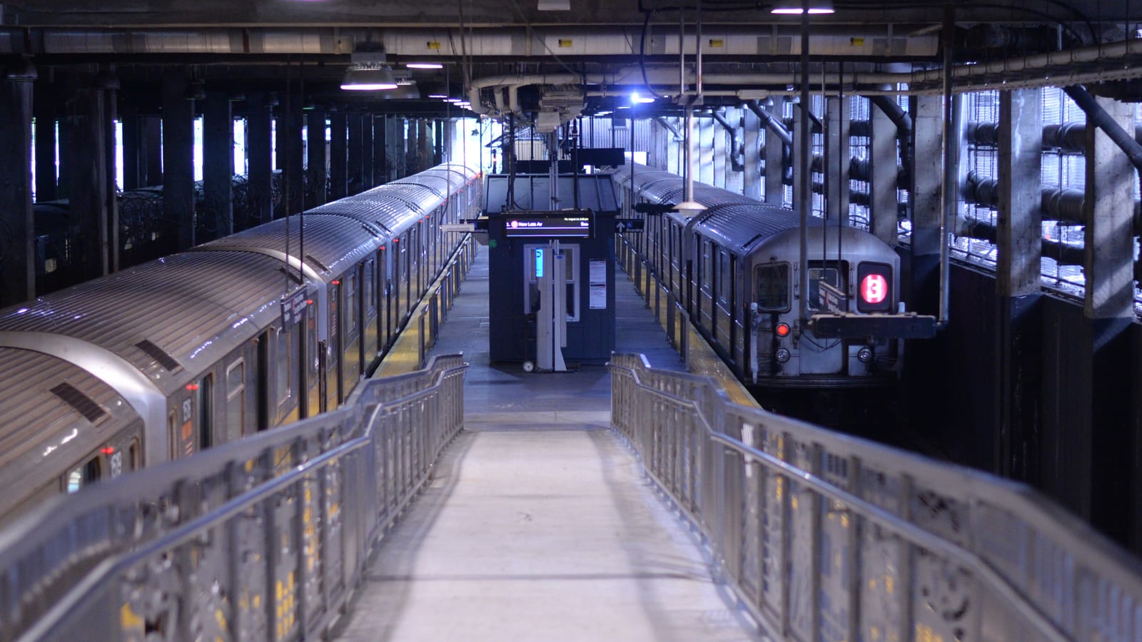 Photograph from a metal ramp looking down onto an indoor subway terminal, with silver trains on two tracks, one showing a red 3 sign, a small blue crew booth between them and pipes and steel beams overhead