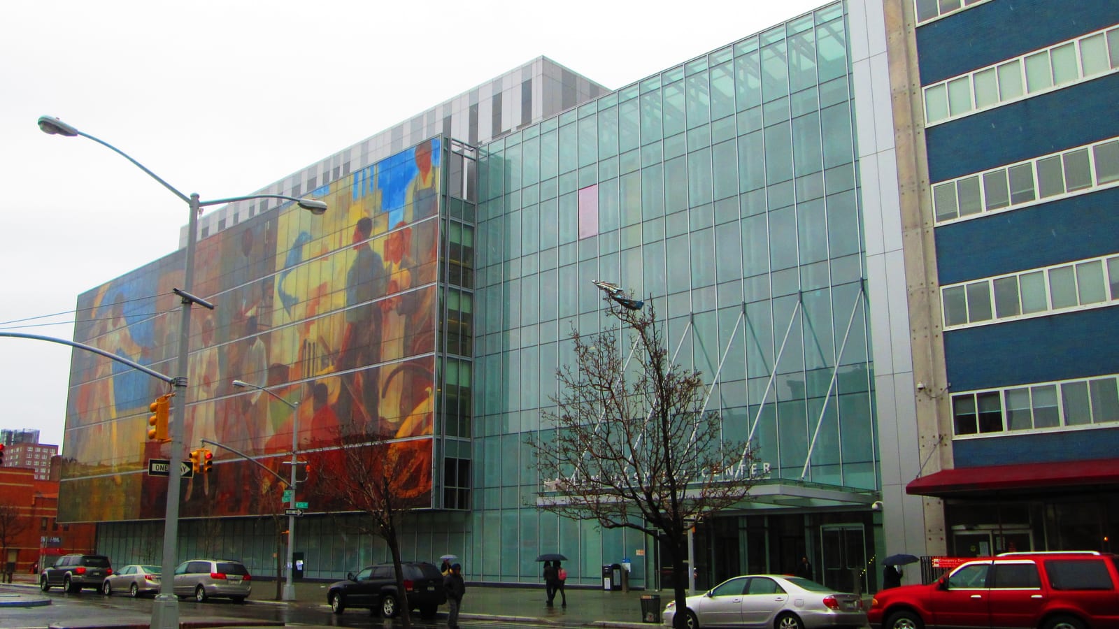Photograph of a large glass hospital building with a colorful painted mural across its left side, a blue and white older wing at right, a bare tree, street lights and parked cars on a wet, cloudy day