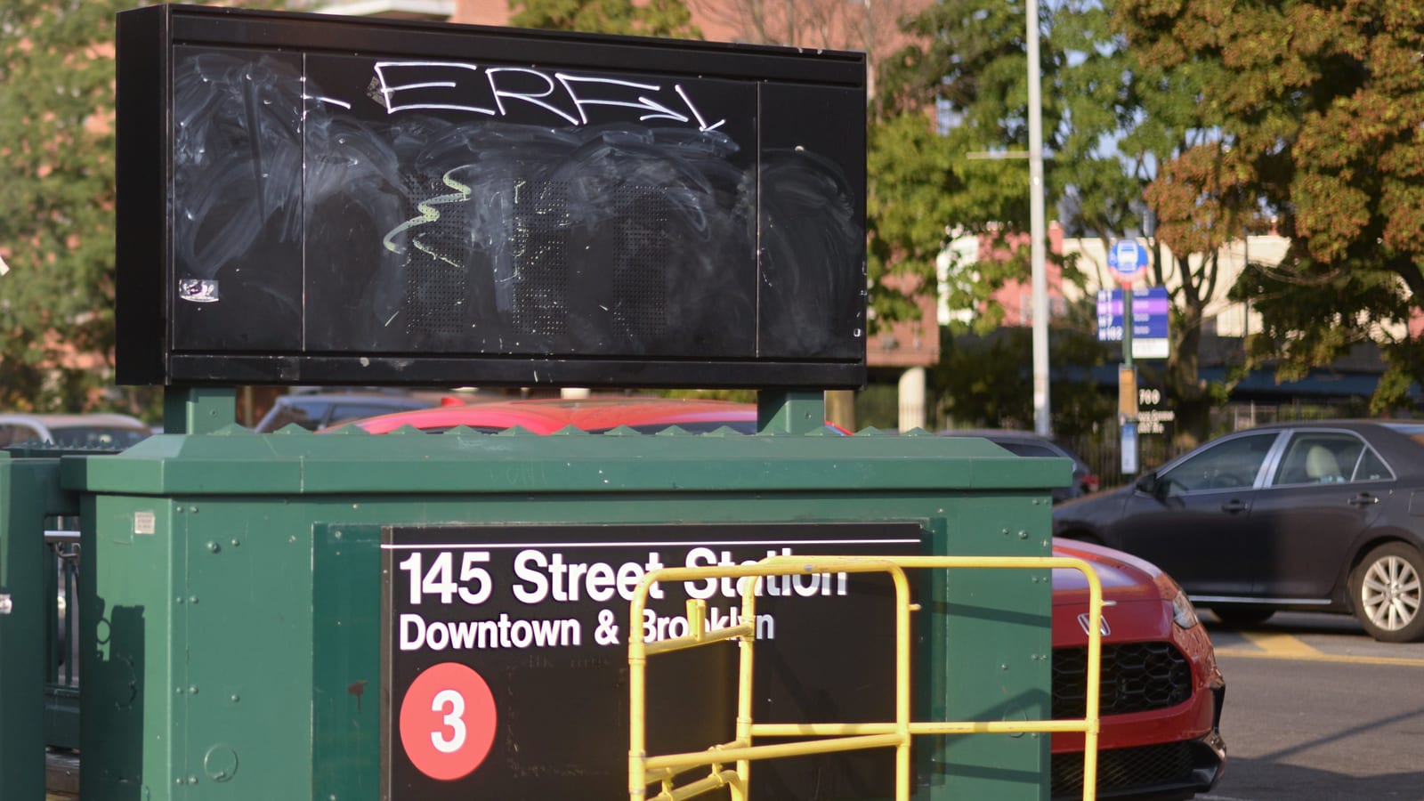 Photograph of a green subway entrance kiosk with a black sign reading 145 Street Station, Downtown and Brooklyn, with a red 3 train symbol, a yellow metal barrier in front, a blank black display board with graffiti above, parked cars at right and trees and a brick apartment building behind
