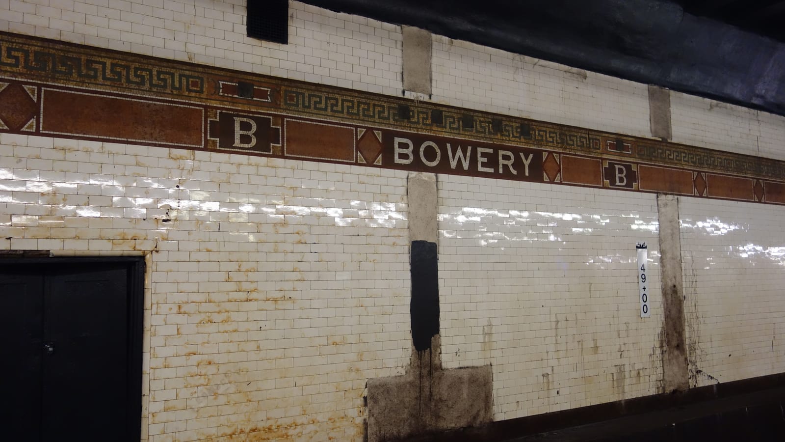 Photograph of a white tiled subway wall with rust stains and a brown mosaic band reading BOWERY between two B tablets, a black door at left and a sign reading 49+00