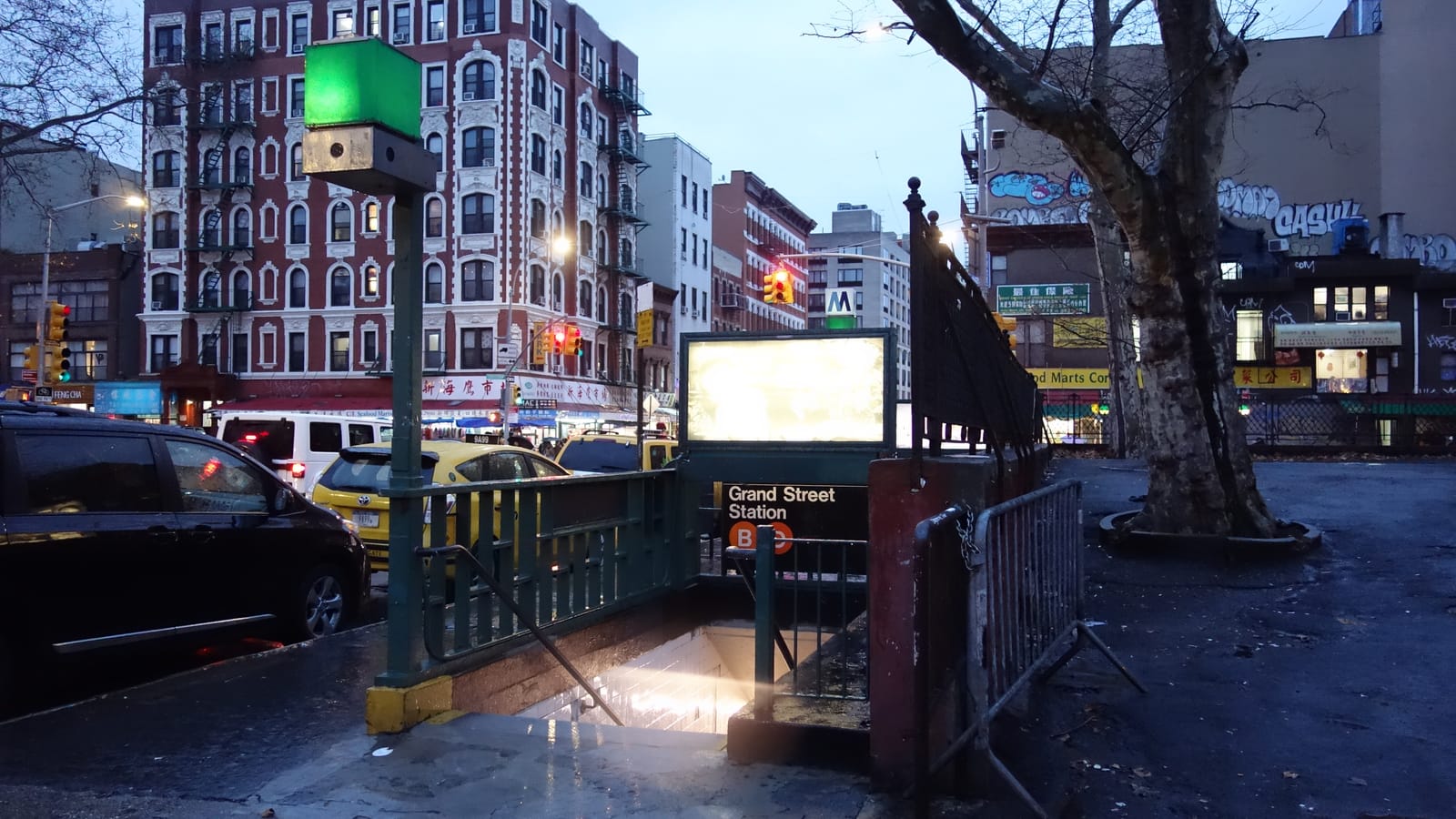 Photograph of a green-railed subway stair entrance with a sign reading Grand Street Station and an orange B line symbol, a green globe lamp, traffic and a yellow taxi on the street, and a red and white apartment building behind at dusk