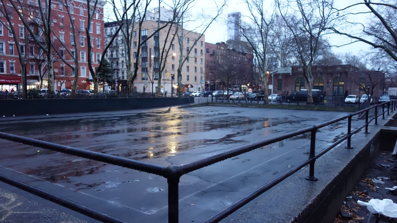 Photograph of a wet, sunken paved court behind black metal railings, with bare trees, parked cars and red brick and tan apartment buildings beyond under a gray evening sky