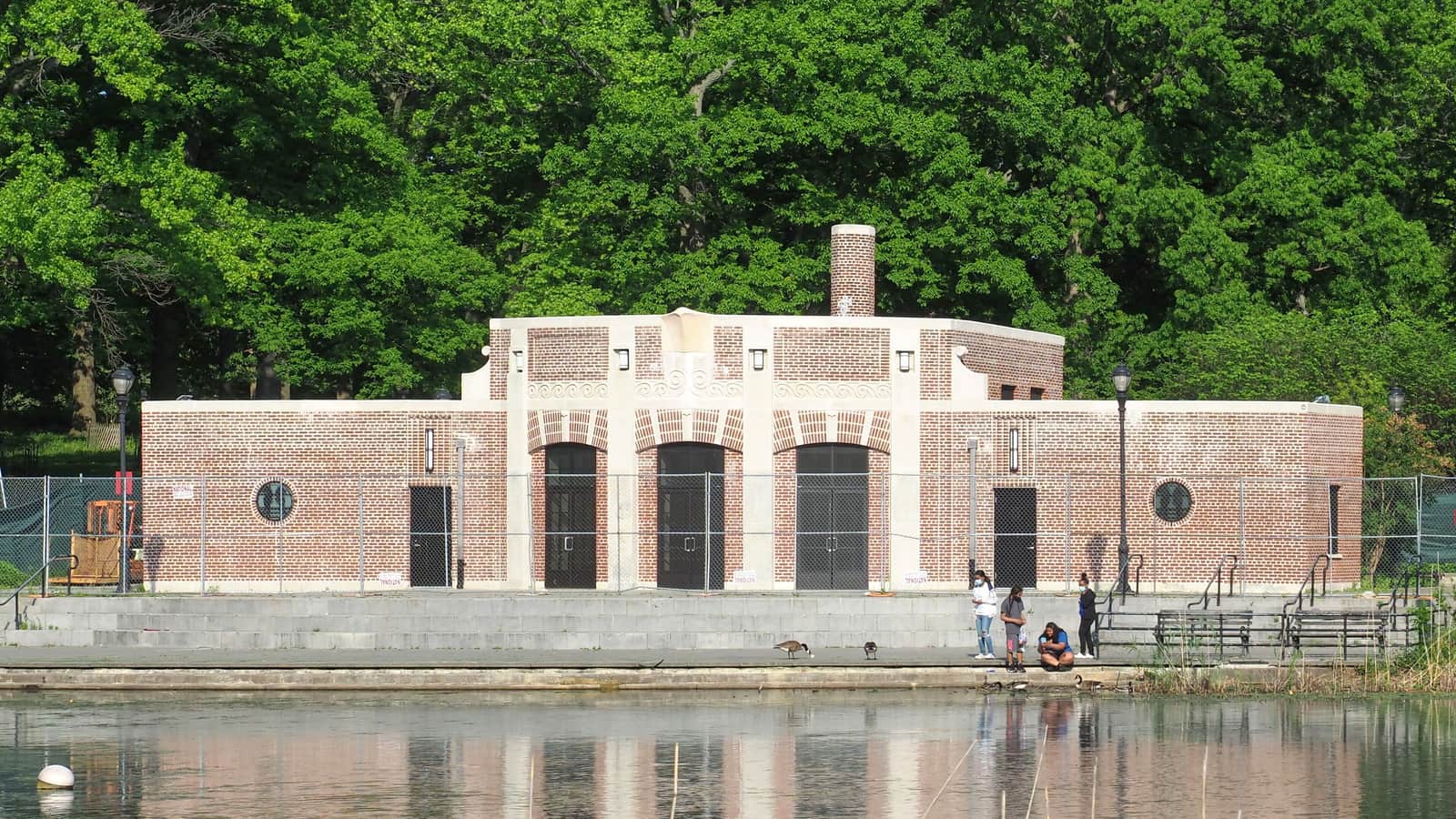 Photograph of a one-story red brick building with three arched doorways, round windows and pale stone trim, behind a temporary wire fence, set on stone steps at the edge of a pond, with tall green trees behind and a few people and geese at the water's edge