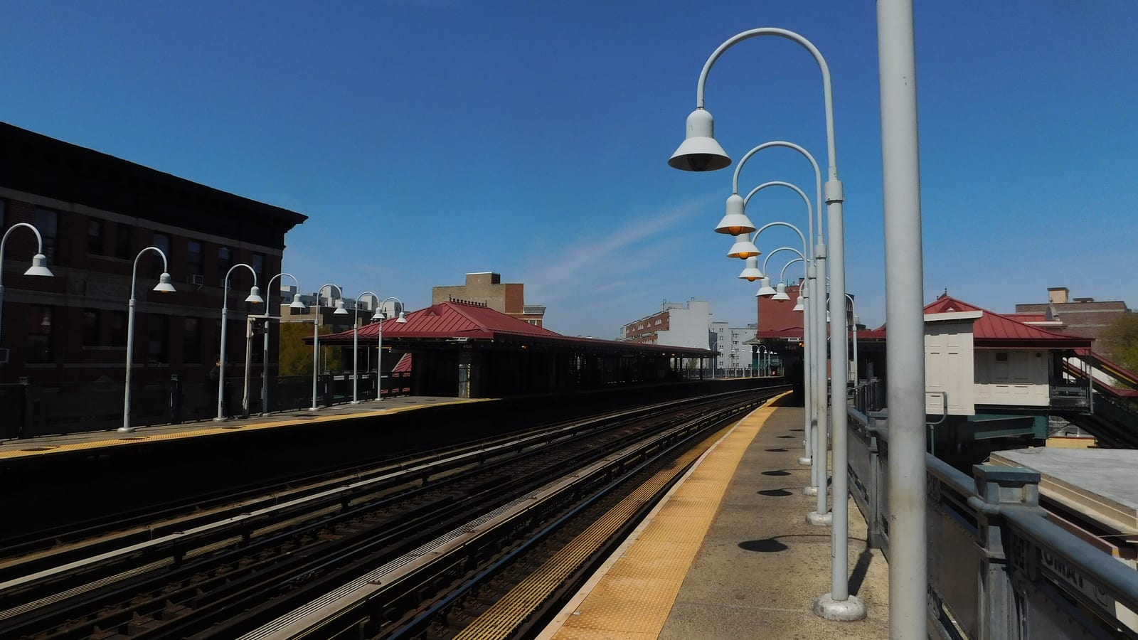 Photograph of an outdoor subway platform under a clear blue sky, with a row of white curved lamp posts along a platform edge with a yellow strip, several tracks, red-roofed platform canopies and a station house at right, and brick apartment buildings behind
