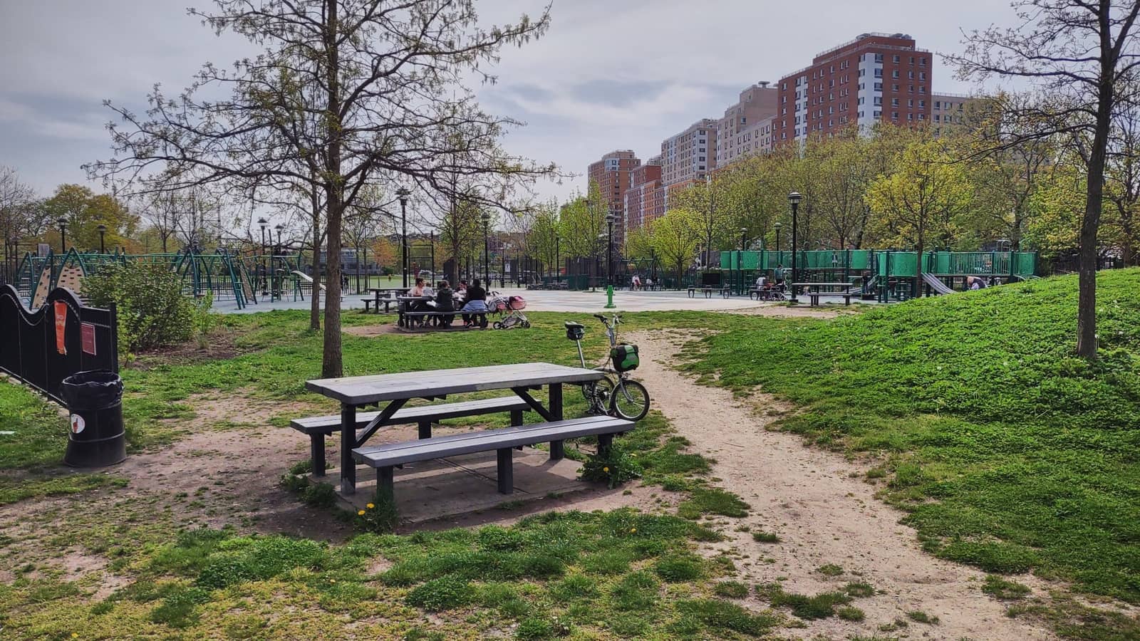 Photograph of a park with a picnic table and a folding bicycle on worn grass in the foreground, a dirt path, a grassy slope at right, green playground equipment and small figures at picnic tables in the middle distance, young trees, and tall brick apartment towers under a cloudy sky