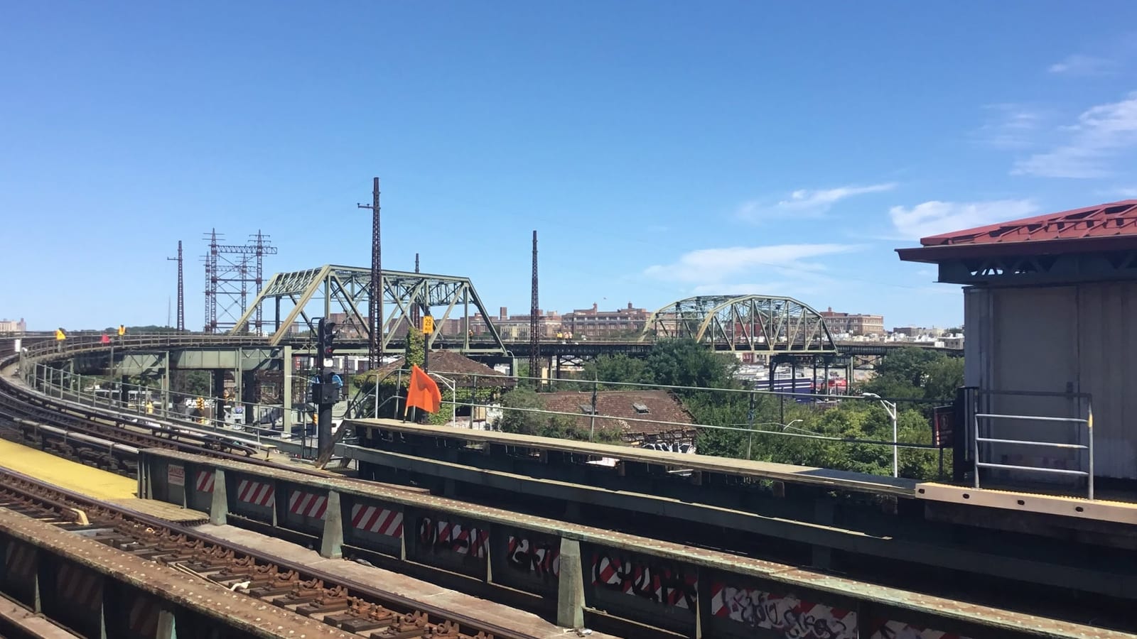 Photograph taken from a subway platform above the street, with tracks and a rusted steel girder in the foreground, a curved track and two green steel truss bridges in the middle distance, an orange flag on a pole, utility towers, trees and brick apartment buildings under a blue sky, and the corner of a red-roofed platform shelter at right