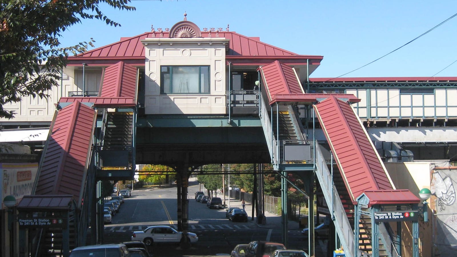 Photograph of an elevated subway station house with a red metal roof and a pale facade above a street, with two covered red stairways down to the sidewalk, green entrance signs reading Freeman St Station for the 2 and 5 trains, the steel structure of the line, and parked cars on a street running downhill under the tracks