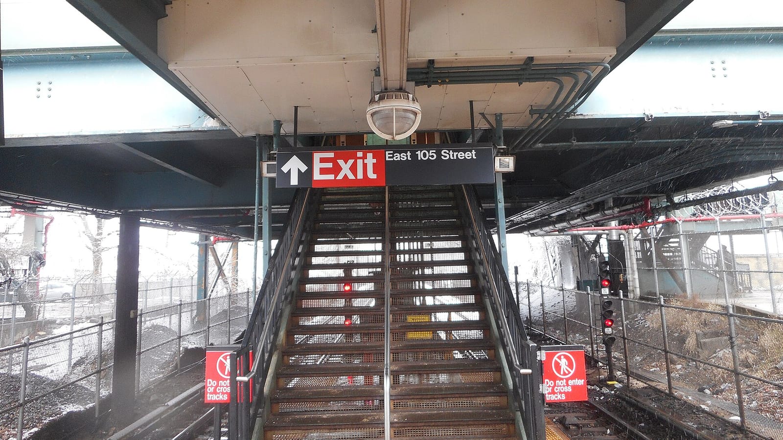 A metal staircase rising from a subway platform to an overhead walkway, under a sign reading Exit, East 105 Street with an up arrow; red Do not enter or cross tracks signs stand at each side of the stairs, tracks and fences run left and right, and light snow is falling