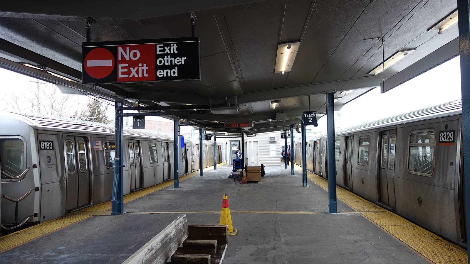 A covered island platform between two silver subway trains numbered 8183 and 8329, with blue columns, yellow edge strips, a traffic cone, a wooden bench in the foreground, Track 1 and Track 2 signs, and a hanging sign reading No Exit, Exit other end