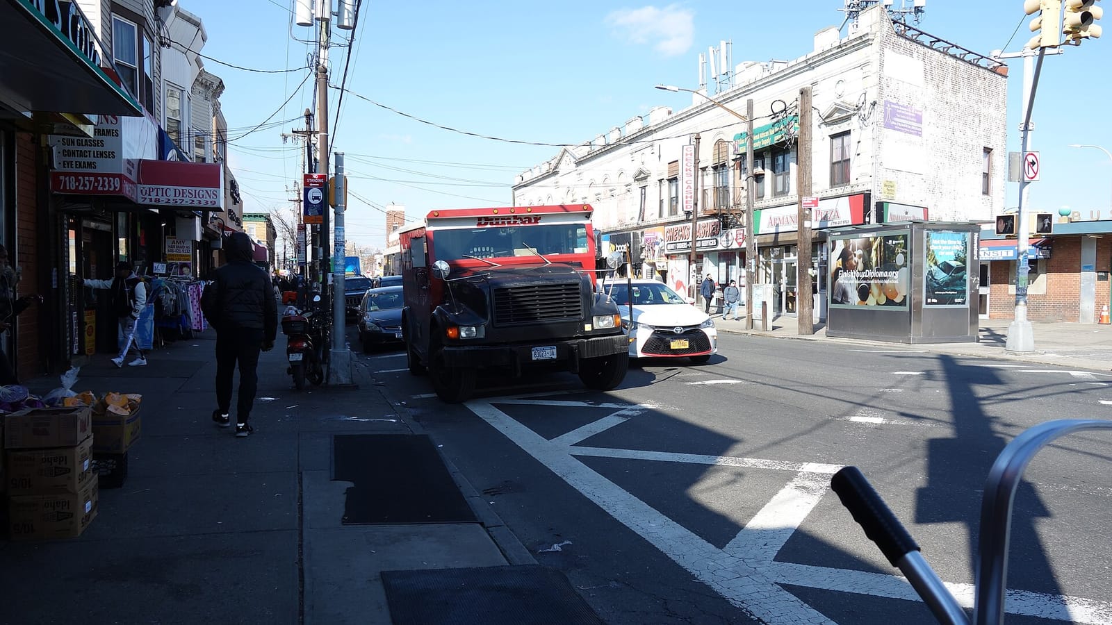 A busy shopping street on a sunny day: a sidewalk at left with produce boxes and clothing racks in front of shops with signs for an optician and a hair braider, a person in a dark jacket walking away, a red and black armored truck and a white taxi stopped at a crosswalk, and two- and three-story buildings with a check-cashing store and a pizzeria across the street