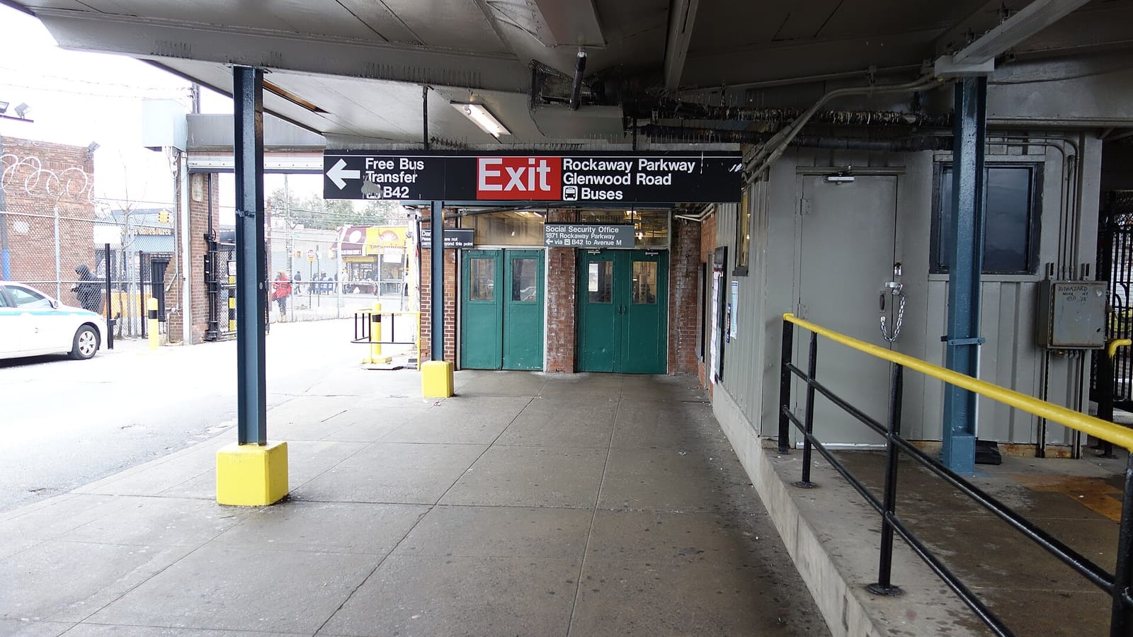 A covered walkway leading to two pairs of green doors under an overhead sign reading Exit, Rockaway Parkway, Glenwood Road, Buses, with an arrow to the left for a free bus transfer to the B42, a yellow-and-black railing at right and a parked white car at left