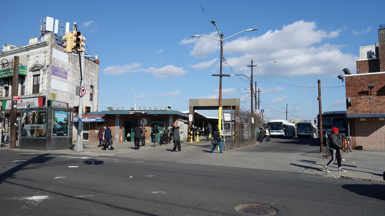 A low brick station house with a sign reading Bus and Subway and a Rockaway Parkway Station sign over two open doorways, people walking on the wide sidewalk in front, a newsstand kiosk with posters at left beside a corner building with a pizzeria, and a fenced bus loop with several city buses at right under a blue sky