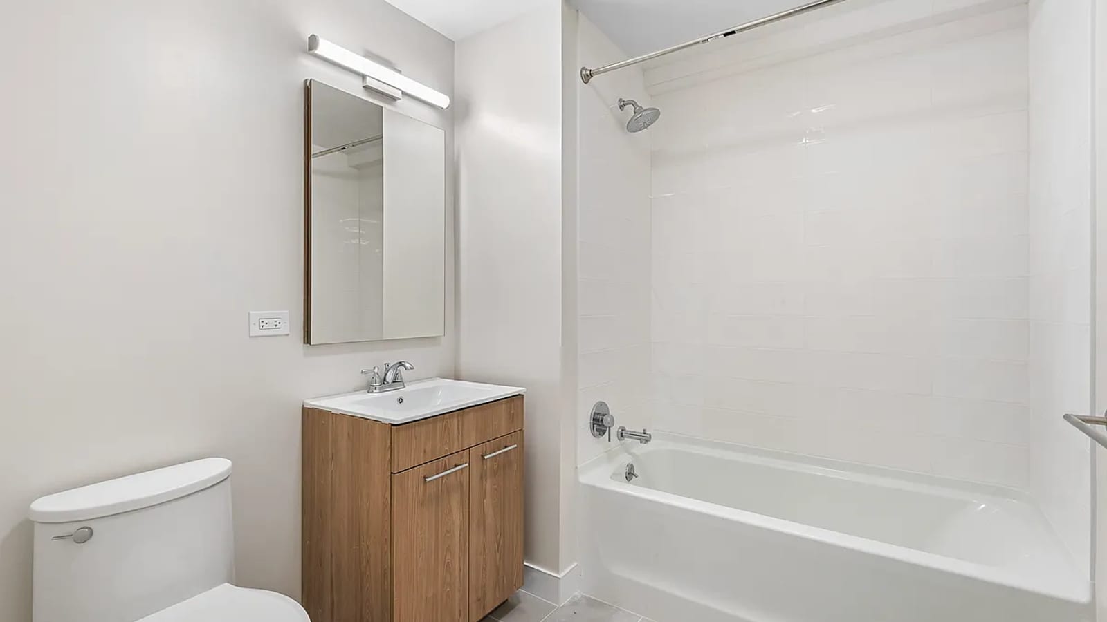 Photograph of a bathroom with a white tub under a tiled shower wall, a wood vanity with a white sink, a mirror cabinet under a bar light, and a toilet