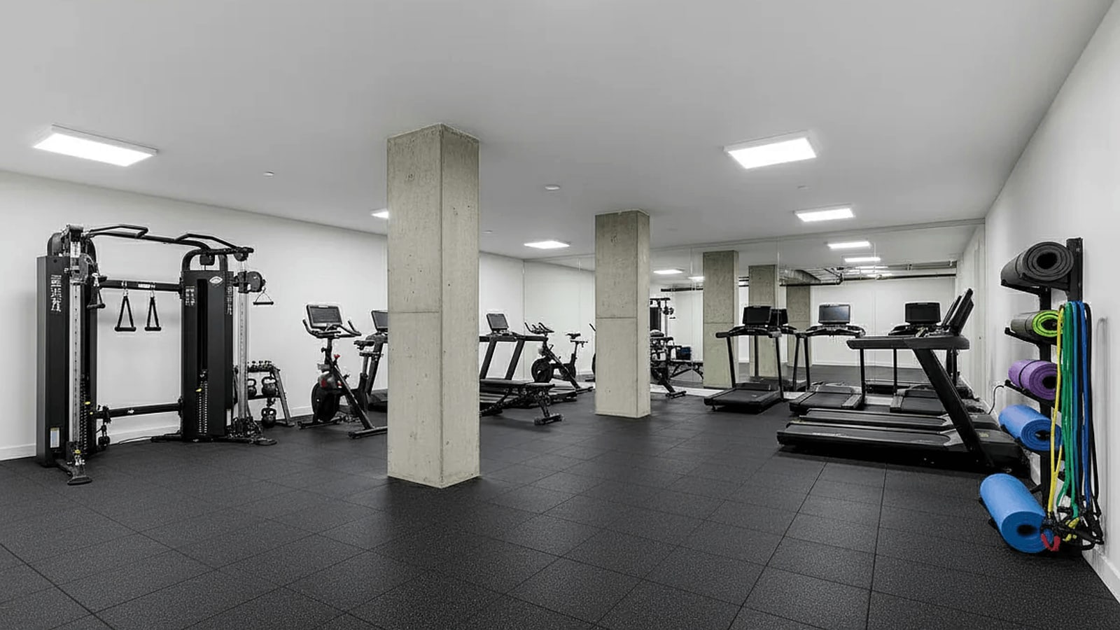 A basement gym with a cable machine, exercise bikes, treadmills and a rack of yoga mats and resistance bands, between concrete columns on black rubber flooring