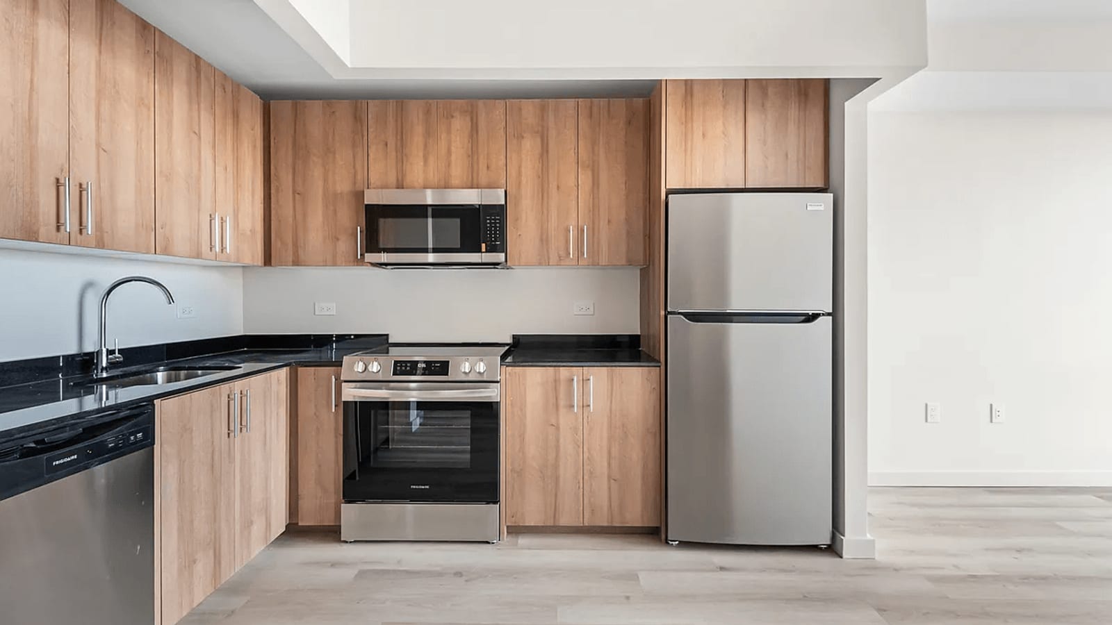 Photograph of an empty kitchen with wood-grain cabinets, black stone counters, a stainless steel dishwasher, a range with a flat cooktop and a microwave above it, a two-door refrigerator and light wood-look flooring