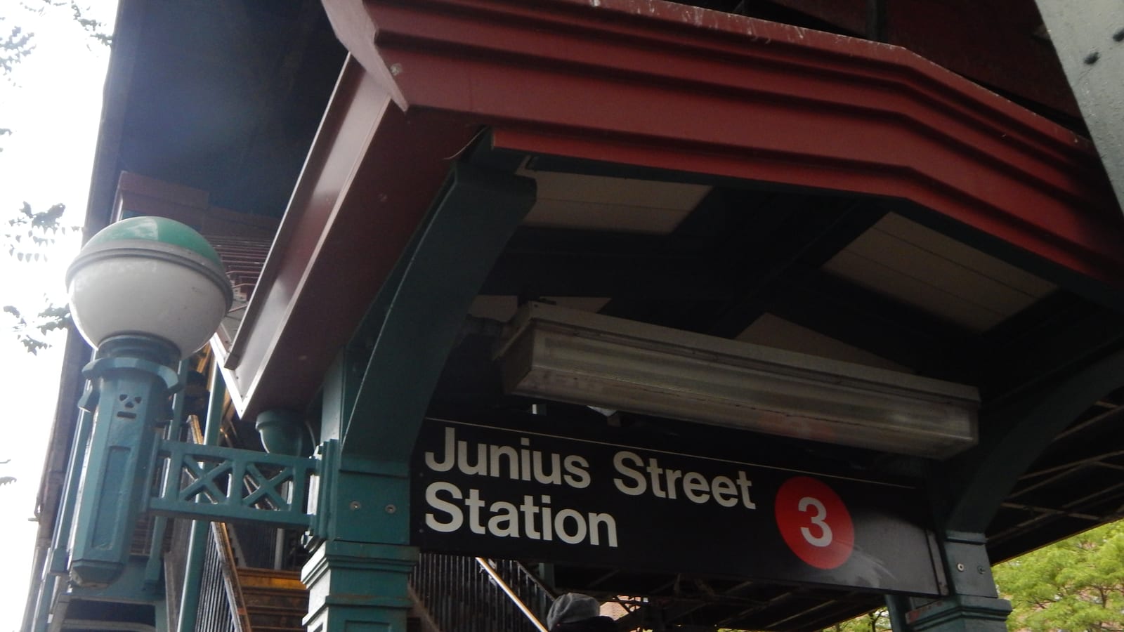 Photograph looking up at a subway stair entrance under an elevated line, with a red-trimmed canopy, a green lamp post with a white globe and a black sign reading Junius Street Station with a red 3 symbol