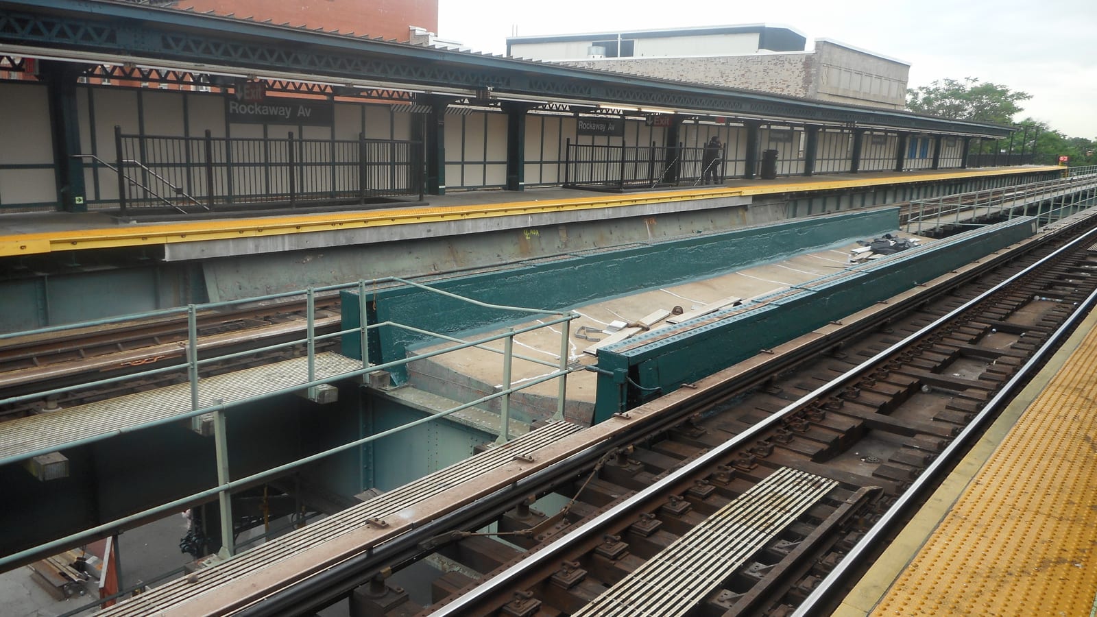 Photograph across the tracks of an elevated station, with a platform under a green steel canopy and signs reading Rockaway Av, a yellow platform edge, a green-painted steel beam and walkway over the street below, rails in the foreground and brick buildings and trees behind